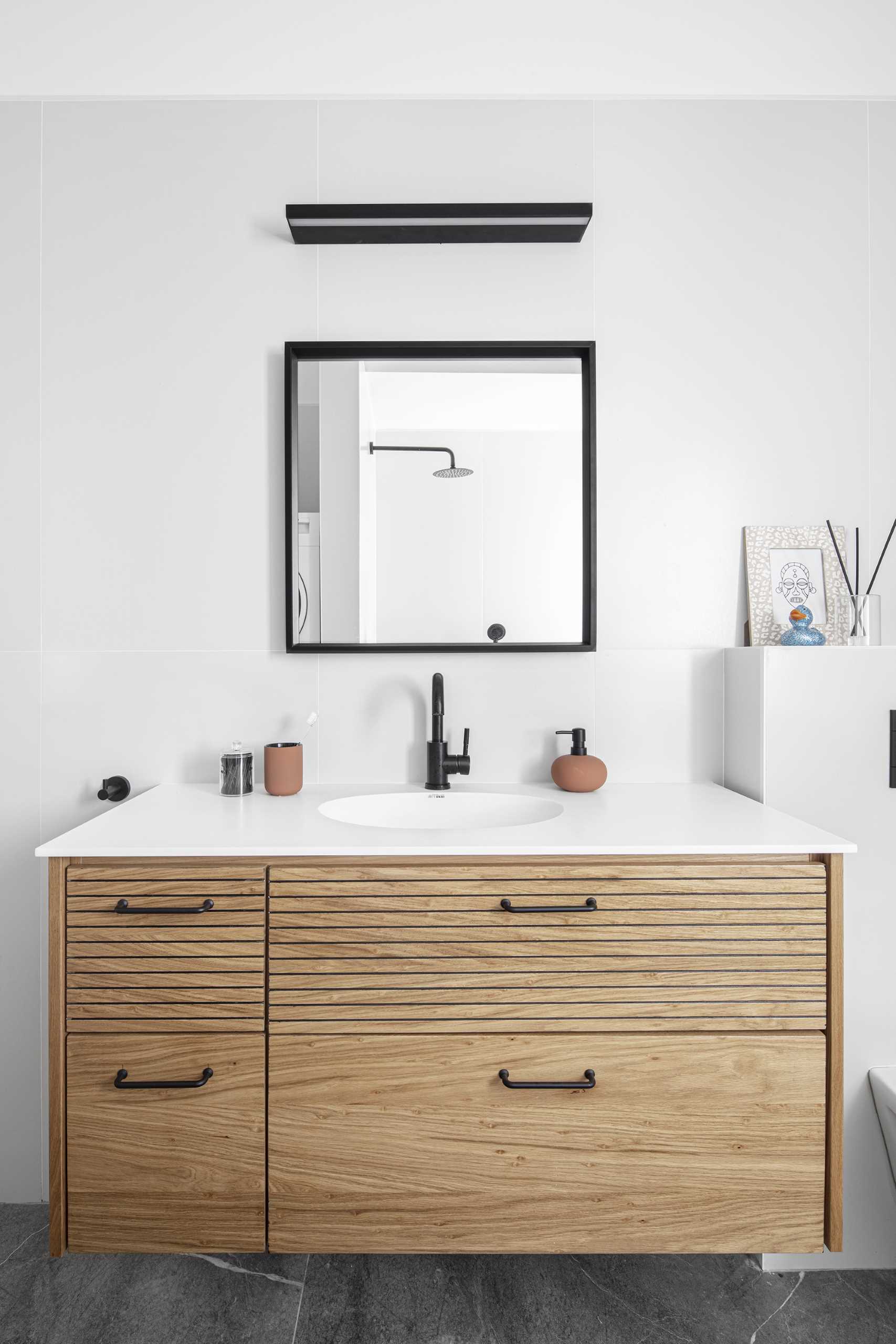 In this modern bathroom, there's a floating wood vanity, a black-framed-square mirror, a pair of wall hooks, large format tiles, and a bath/shower combo. Hidden from view are the laundry machines.