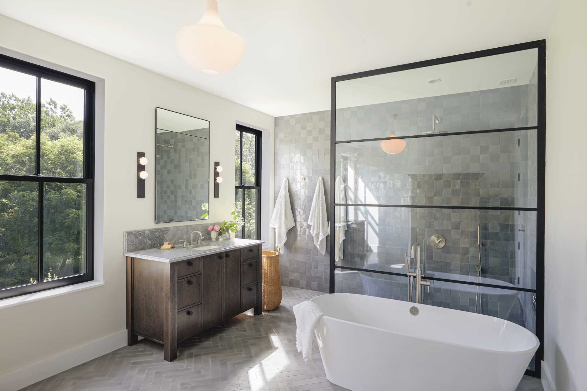 In this modern bathroom, there's square grey tiles covering the wall, and a black framed glass screen separates the freestanding bathtub from the shower.