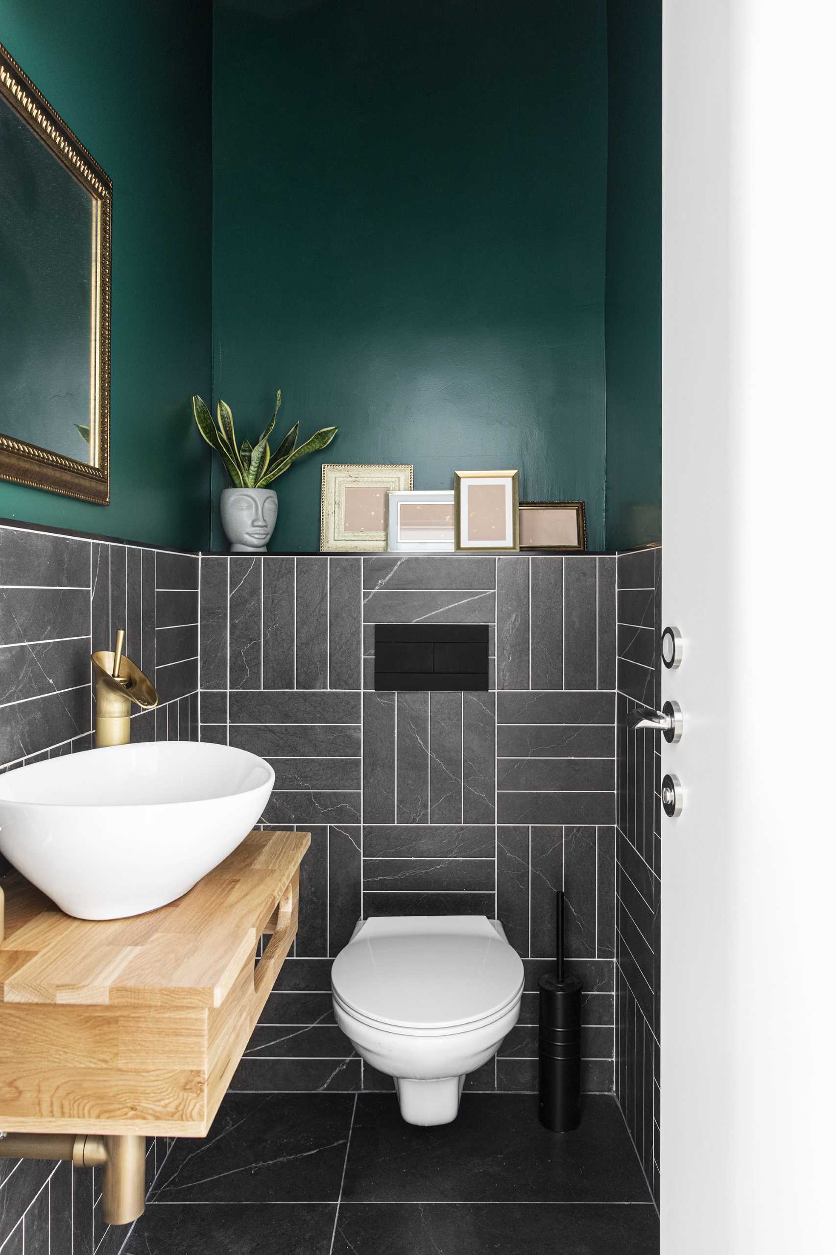 Este lavabo moderno tem paredes verdes arrojadas, azulejos cinza escuro e um espelho de moldura dourada com itens de decoração correspondentes.