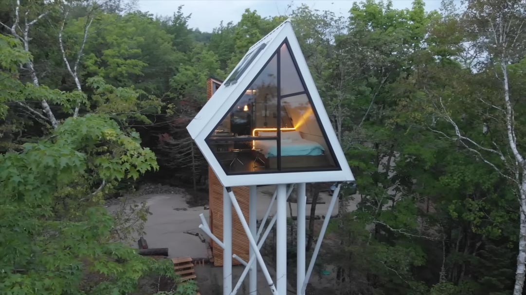 A small cabin is elevated among the trees and is accessed via an enclosed spiral staircase and a bridge.