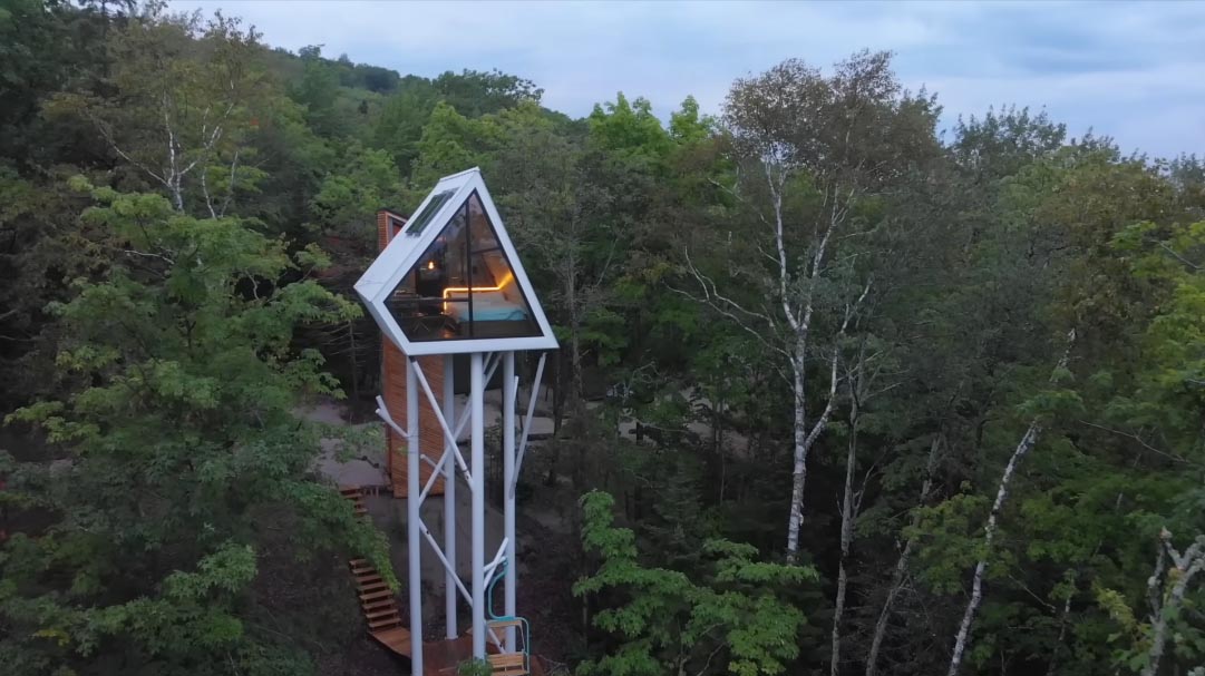 A small cabin is elevated among the trees and is accessed via an enclosed spiral staircase and a bridge.