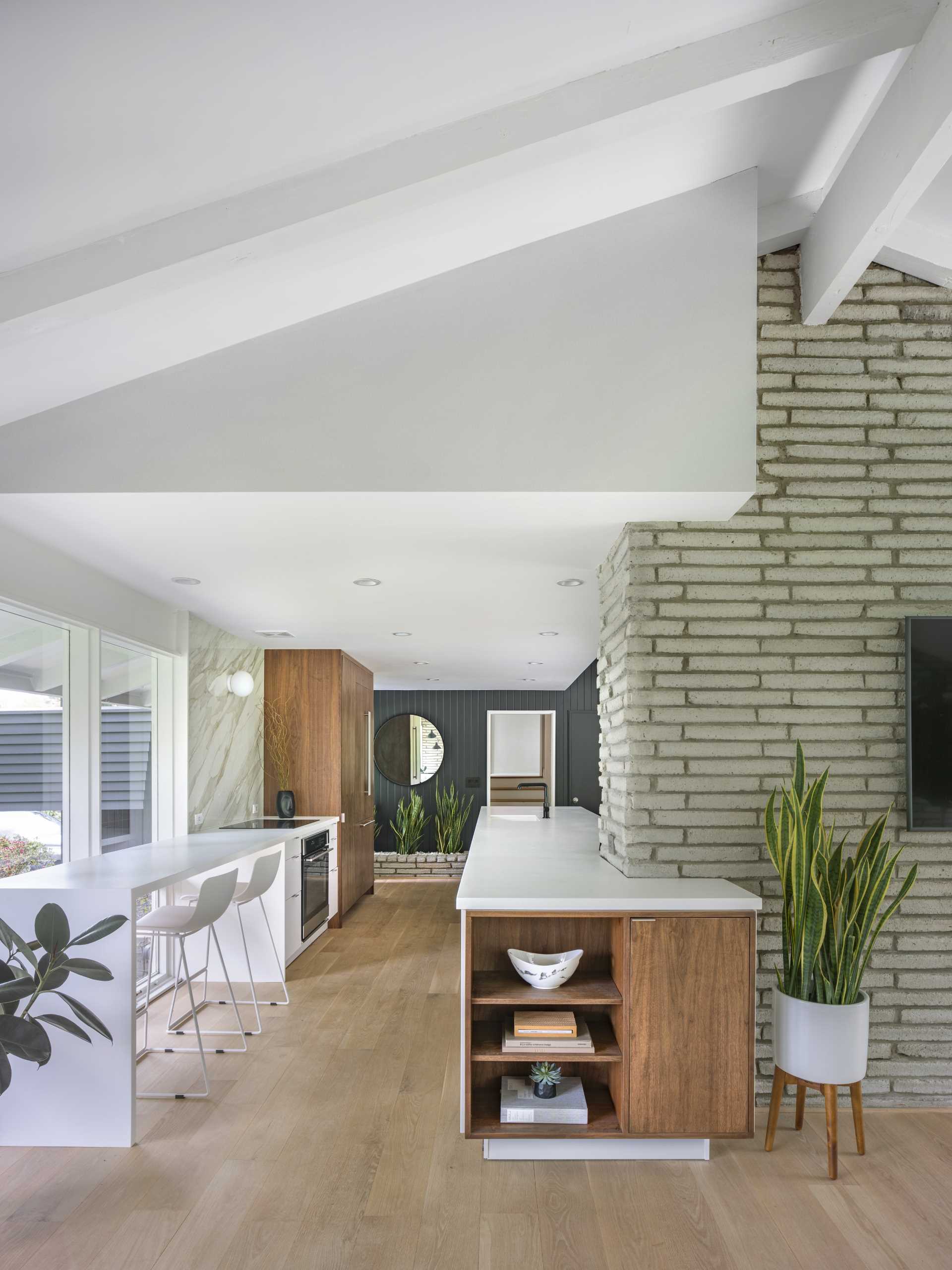 The new kitchen of this renovated mid-century modern home, is bright and open with long white countertops, wood cabinets, and a seating area by the windows.