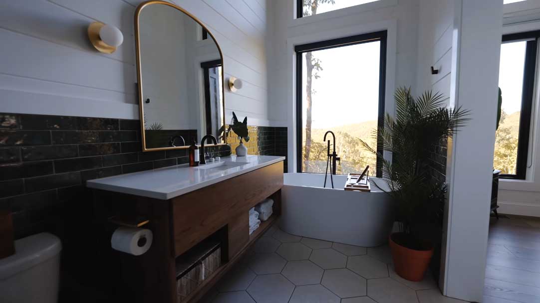 This modern bathroom includes a deep freestanding bathtub by the window, hexagon floor tiles, and a wood vanity with white counter.