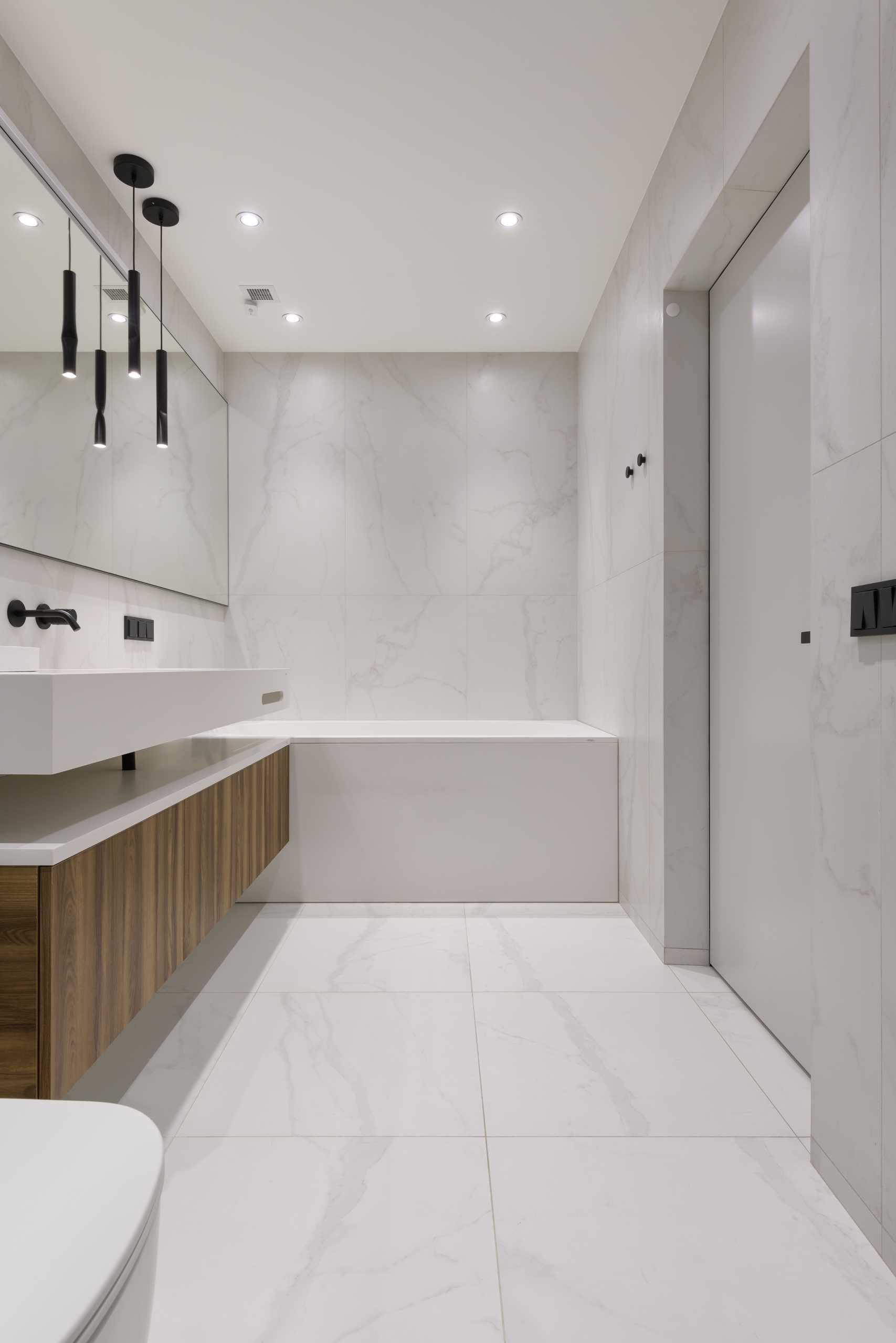 A modern bathroom includes a floating wood and white vanity, a bathtub that perfectly fits between the walls, and a walk-in shower.