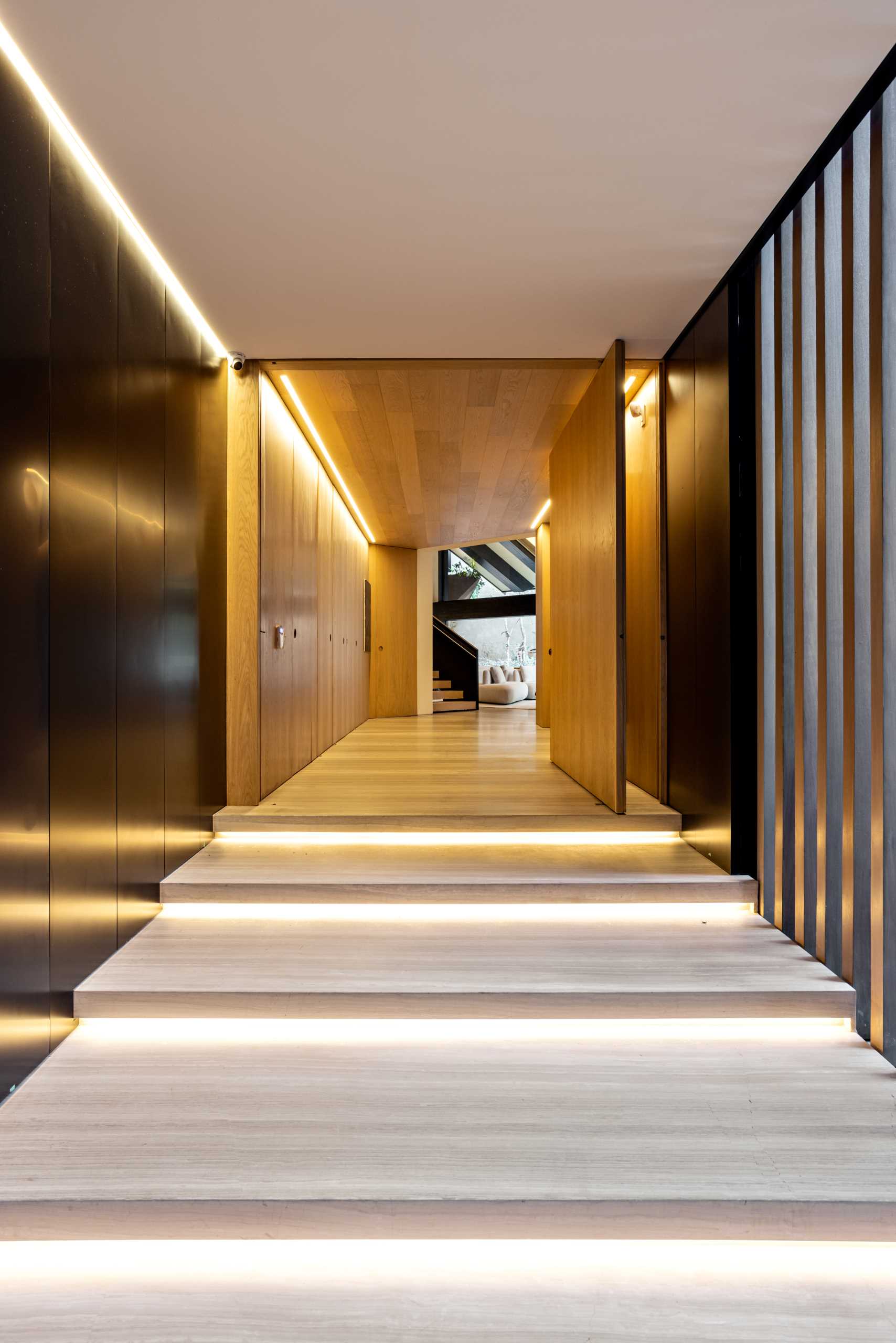 A large pivoting wood front door welcomes people to this modern home. The entryway is lined with wood cabinets and lighting.