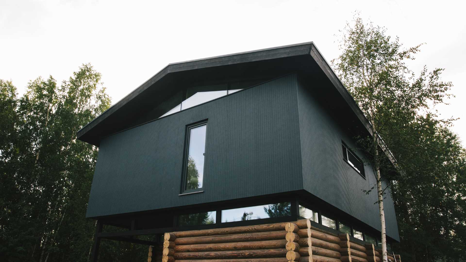 A modern log home with a modern black second floor and roof, and a band of windows.