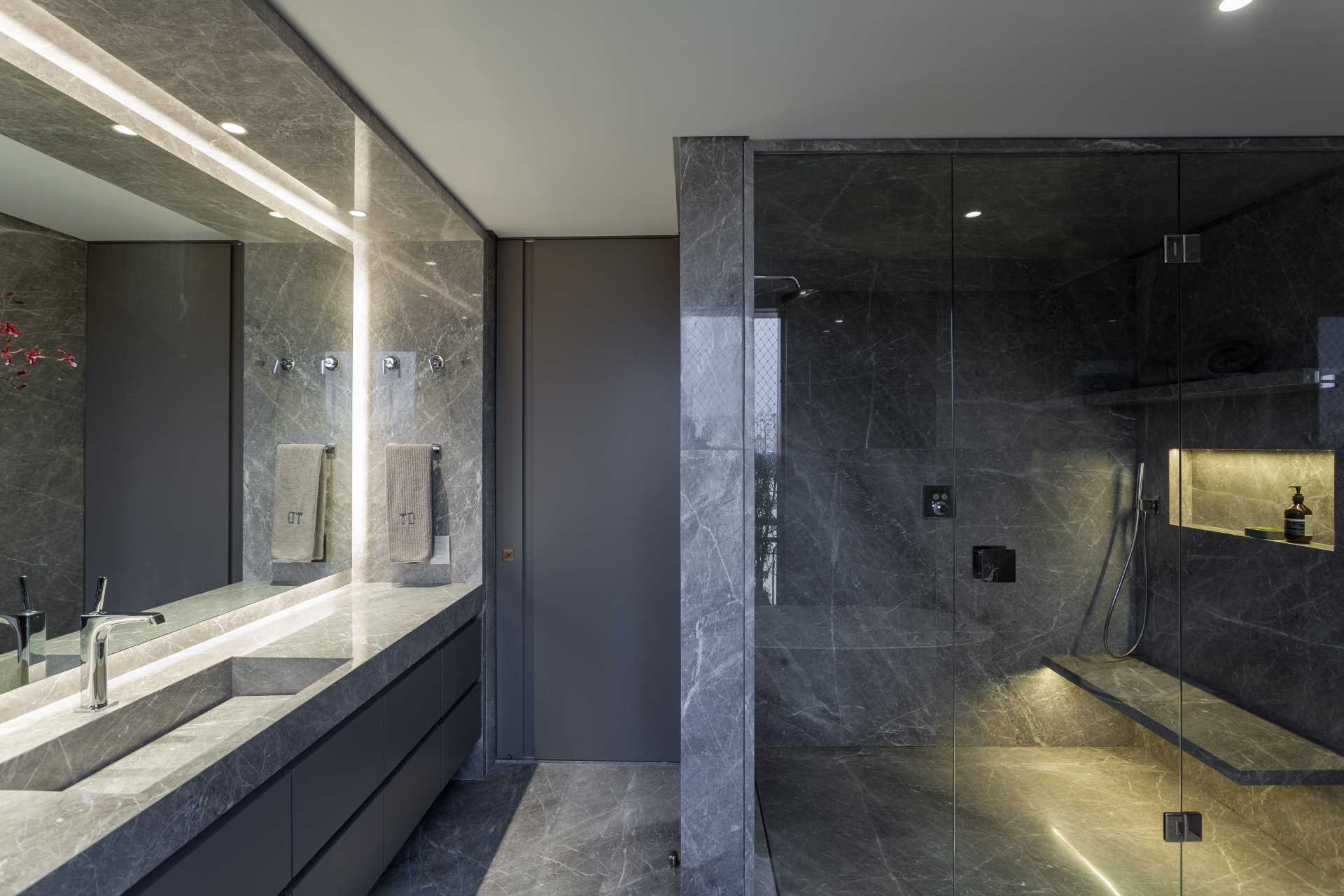 This modern bathroom includes a vanity with built-in sink, a walk-in shower with a bench and shower niche, as well as a freestanding white bathtub by the window.