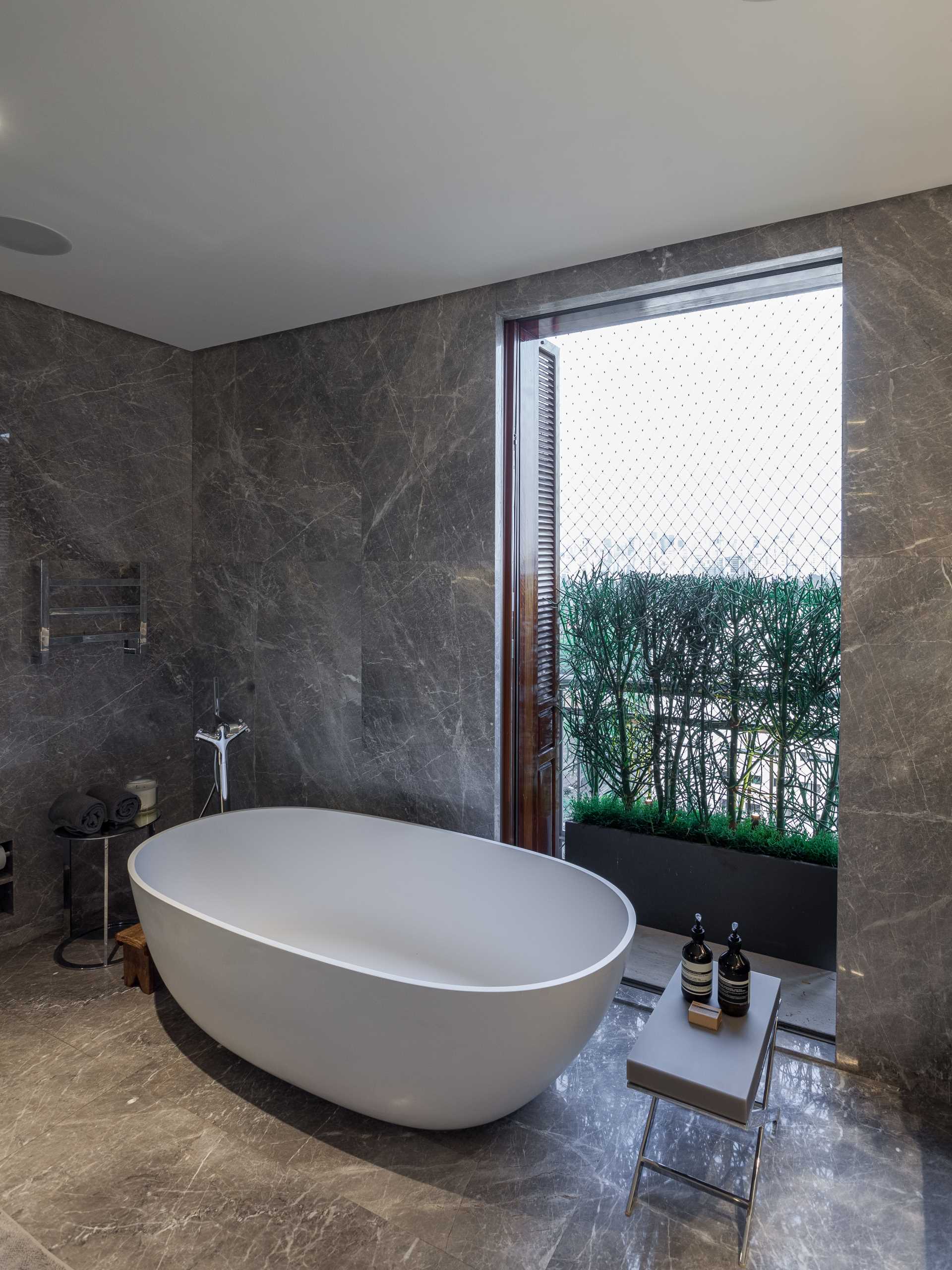 This modern bathroom includes a vanity with built-in sink, a walk-in shower with a bench and shower niche, as well as a freestanding white bathtub by the window.