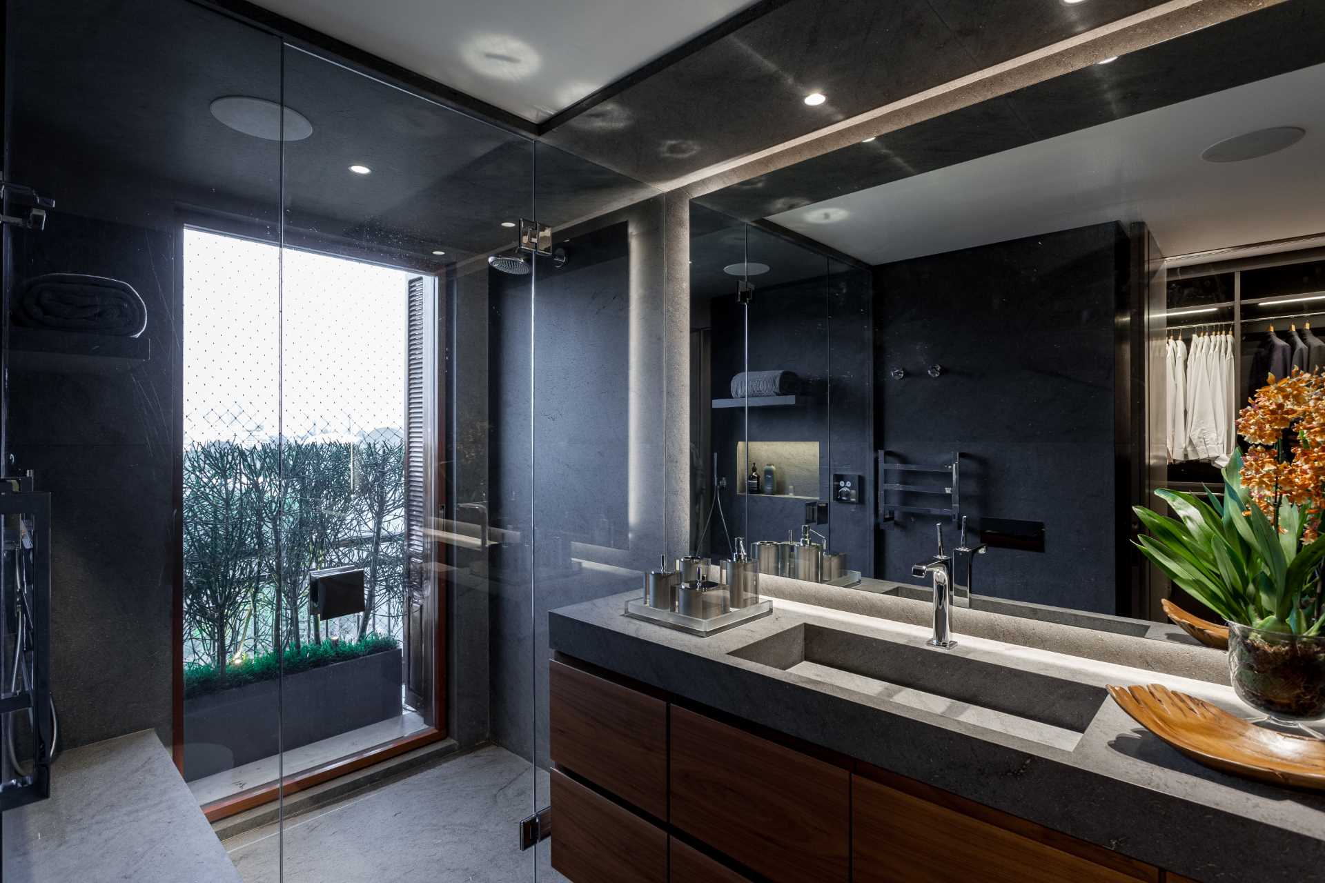 این حمام مدرن شامل دوش شیشه ای در کنار پنجره، روشویی چوبی تیره و یک آینه بزرگ است.