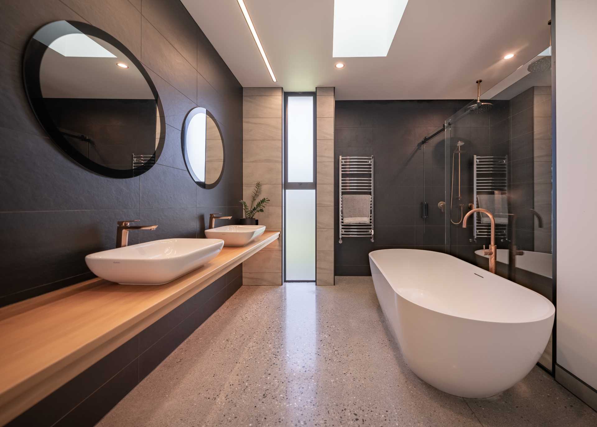 In this modern bathroom, there's a double vanity made from wood, large wall tiles, round mirrors, a freestanding bathtub, and a shower with a glass screen and rainfall showerhead.