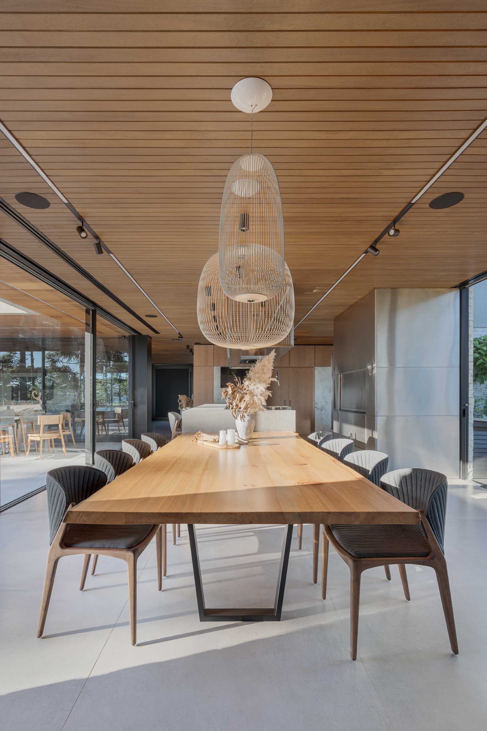 A modern dining area with a long wood table connected to the kitchen island. Dark grey chairs provide a contrasting element, while the wall at the end of the table is dedicated to a pair of glass enclosed wine cellars.