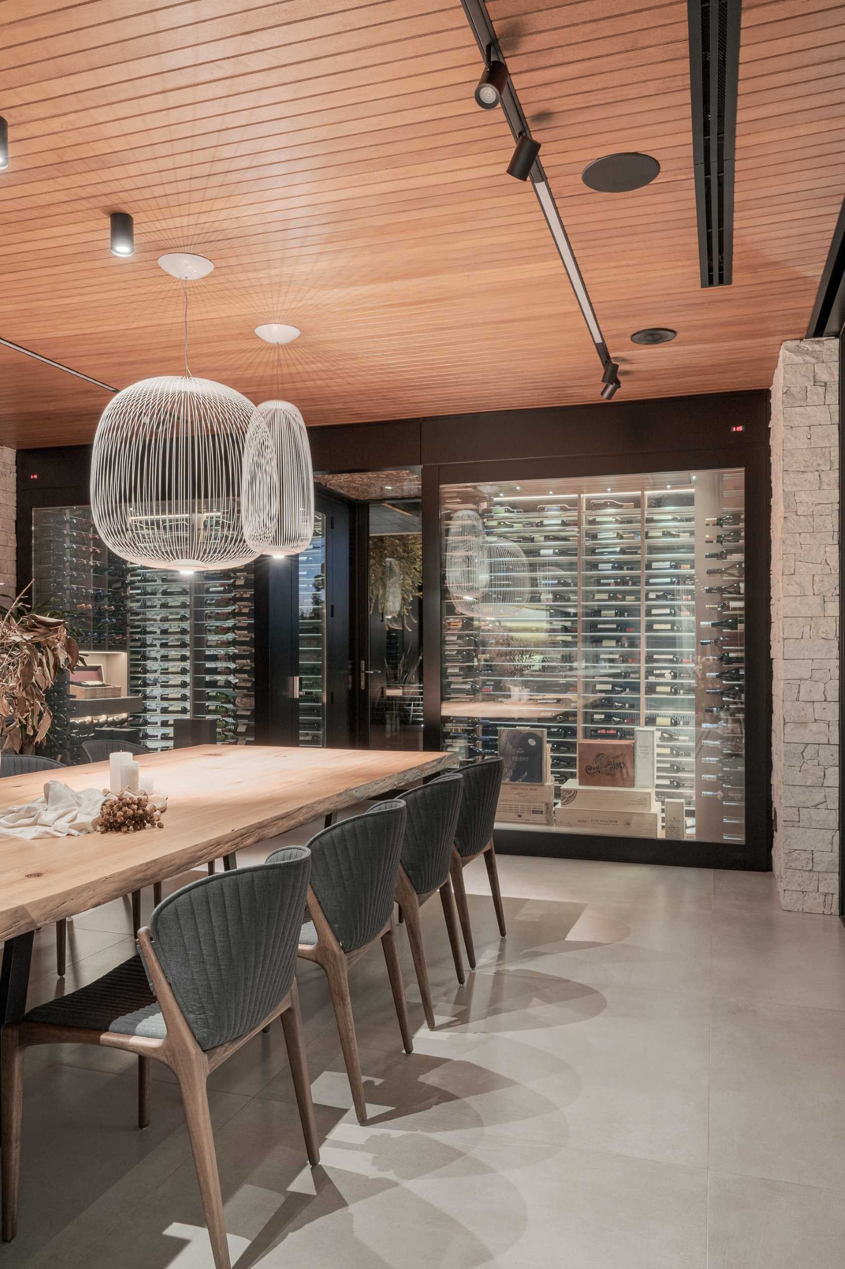 A modern dining area with a long wood table connected to the kitchen island. Dark grey chairs provide a contrasting element, while the wall at the end of the table is dedicated to a pair of glass enclosed wine cellars.