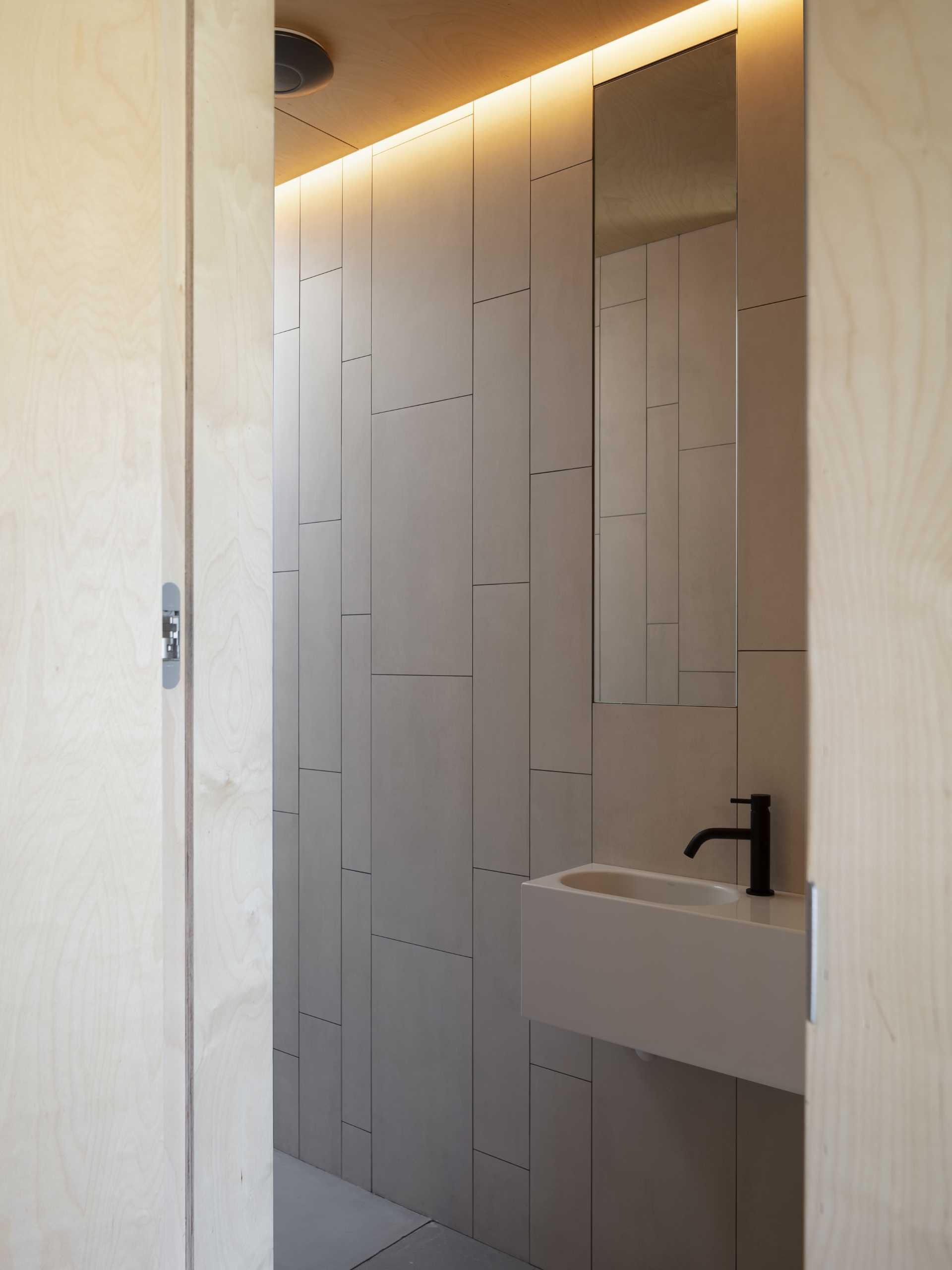 در این حمام مدرن، دیوارها با کاشی پوشانده شده اند، در حالی که یک آینه بلند عمودی چشم را به سمت بالا به نور پنهان می کشاند.