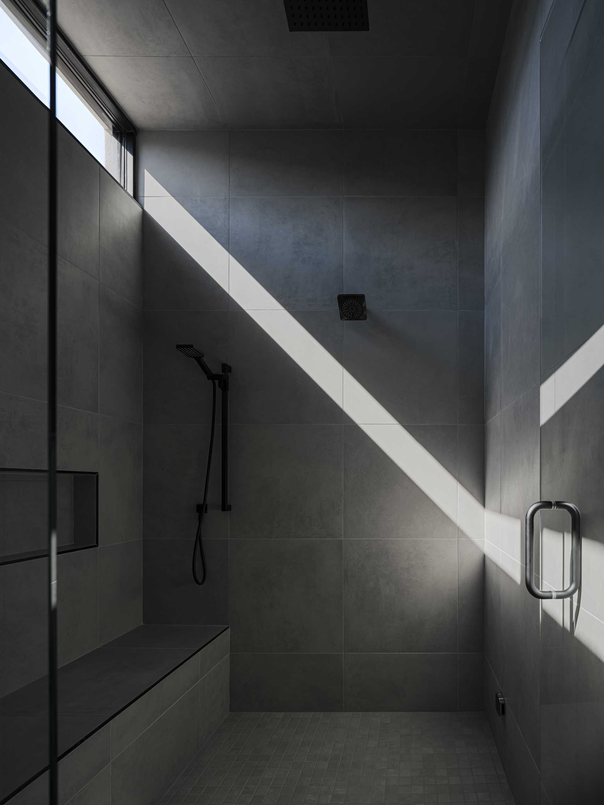 In this dark grey shower, the walls, ceiling, and bench are covered in large tiles, while a horizontal window provides a streak of sunlight.