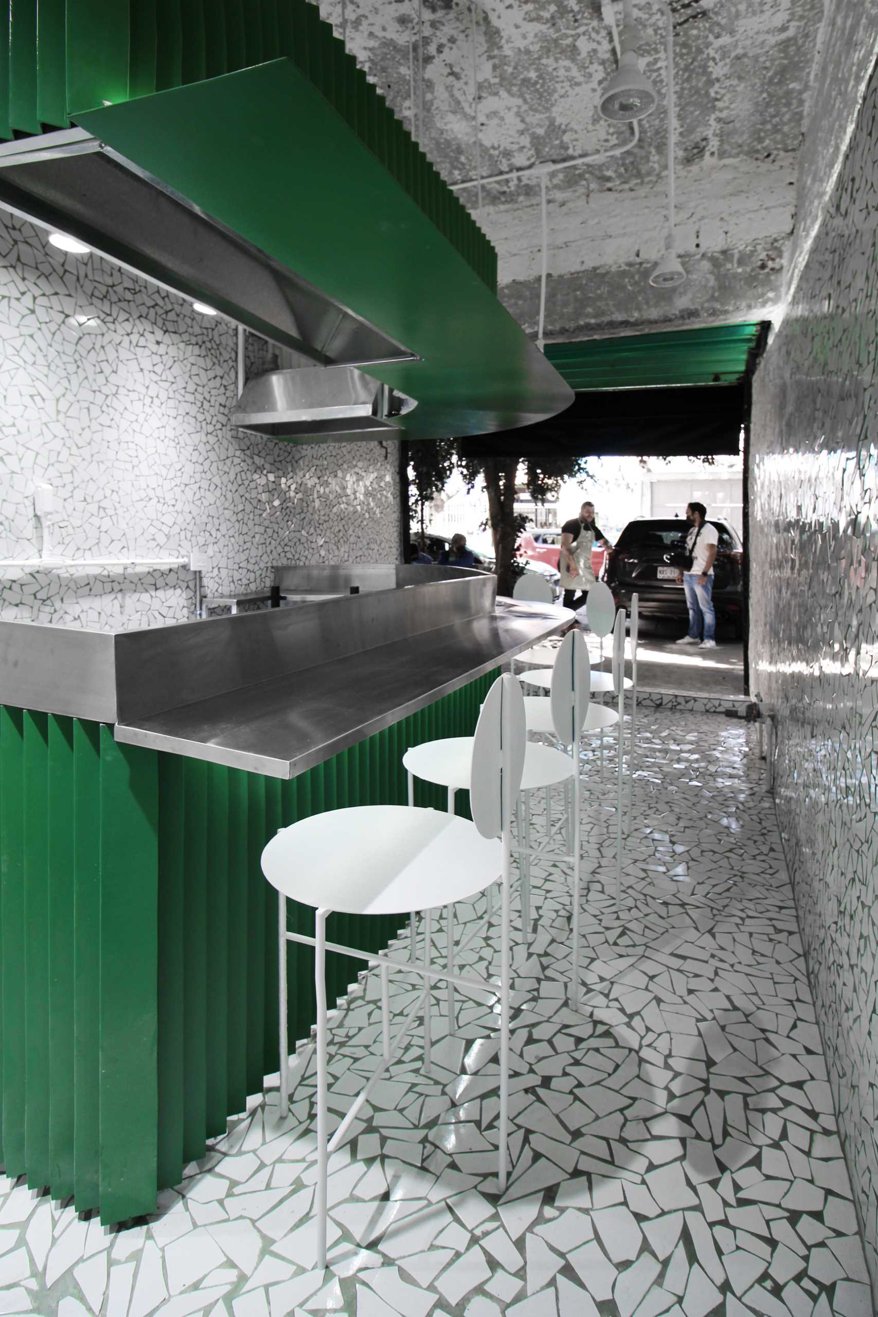 A small taco shop (taqueria) that features broken tile mosaic walls and floor, and a painted green metal accent with white stools.