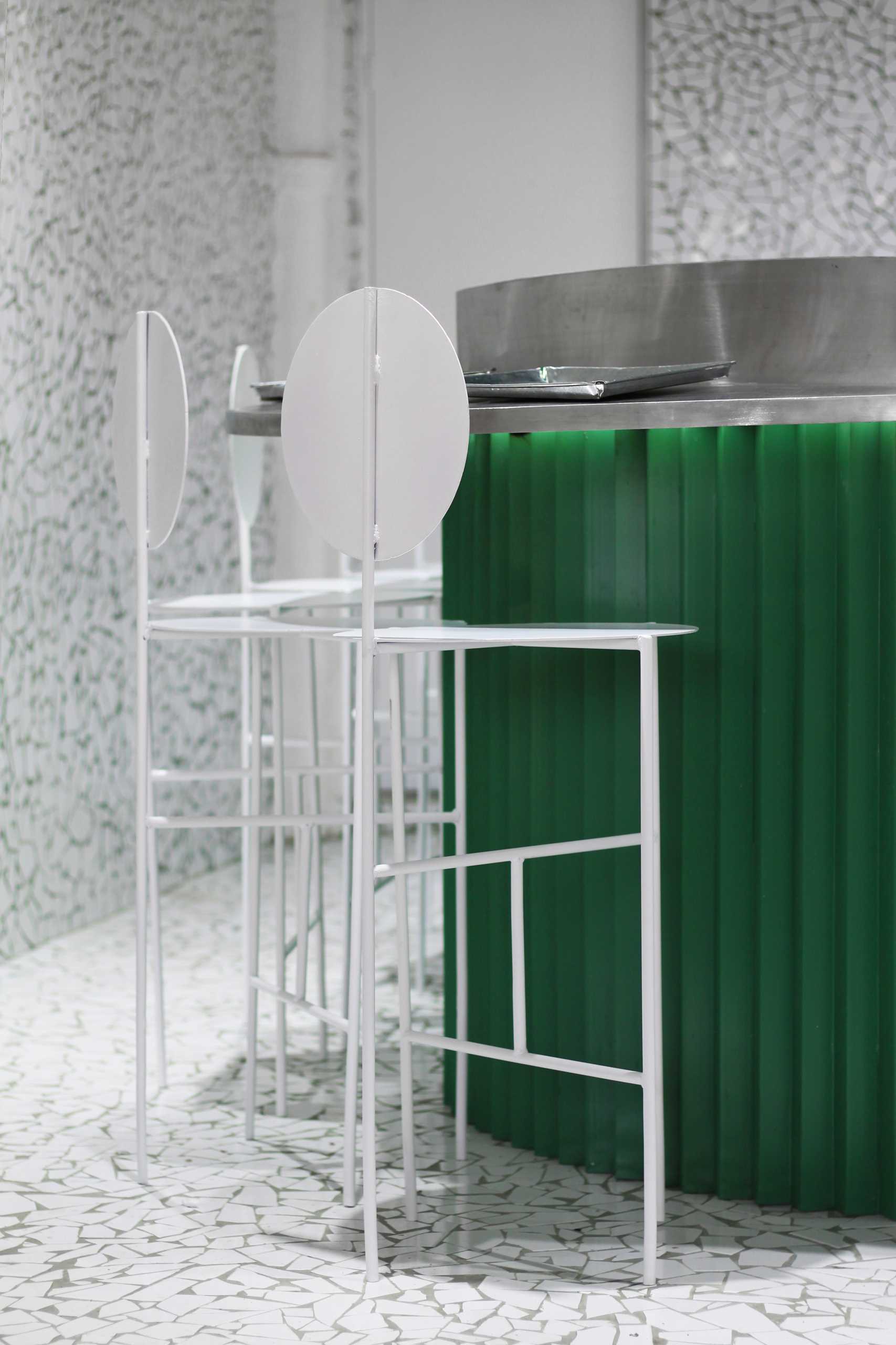 A small taco shop (taqueria) that features broken tile mosaic walls and floor, and a painted green metal counter with white stools.