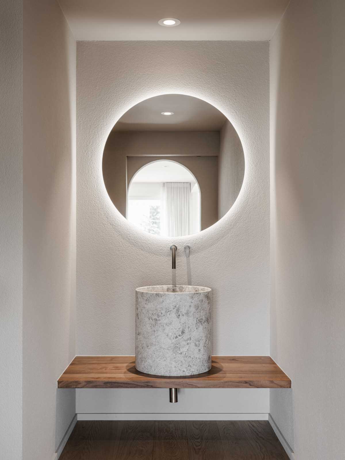 A round back-lit mirror is mounting above a stone sink.