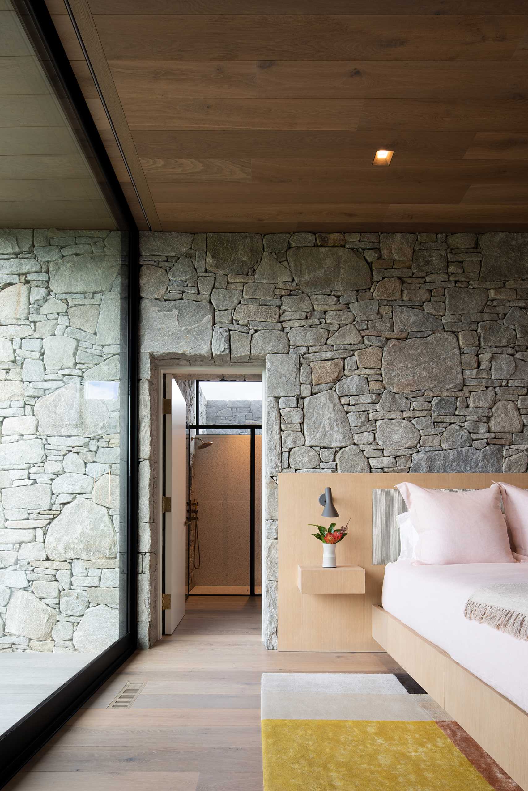 A modern bedroom with stone walls, wood ceiling, and large windows.