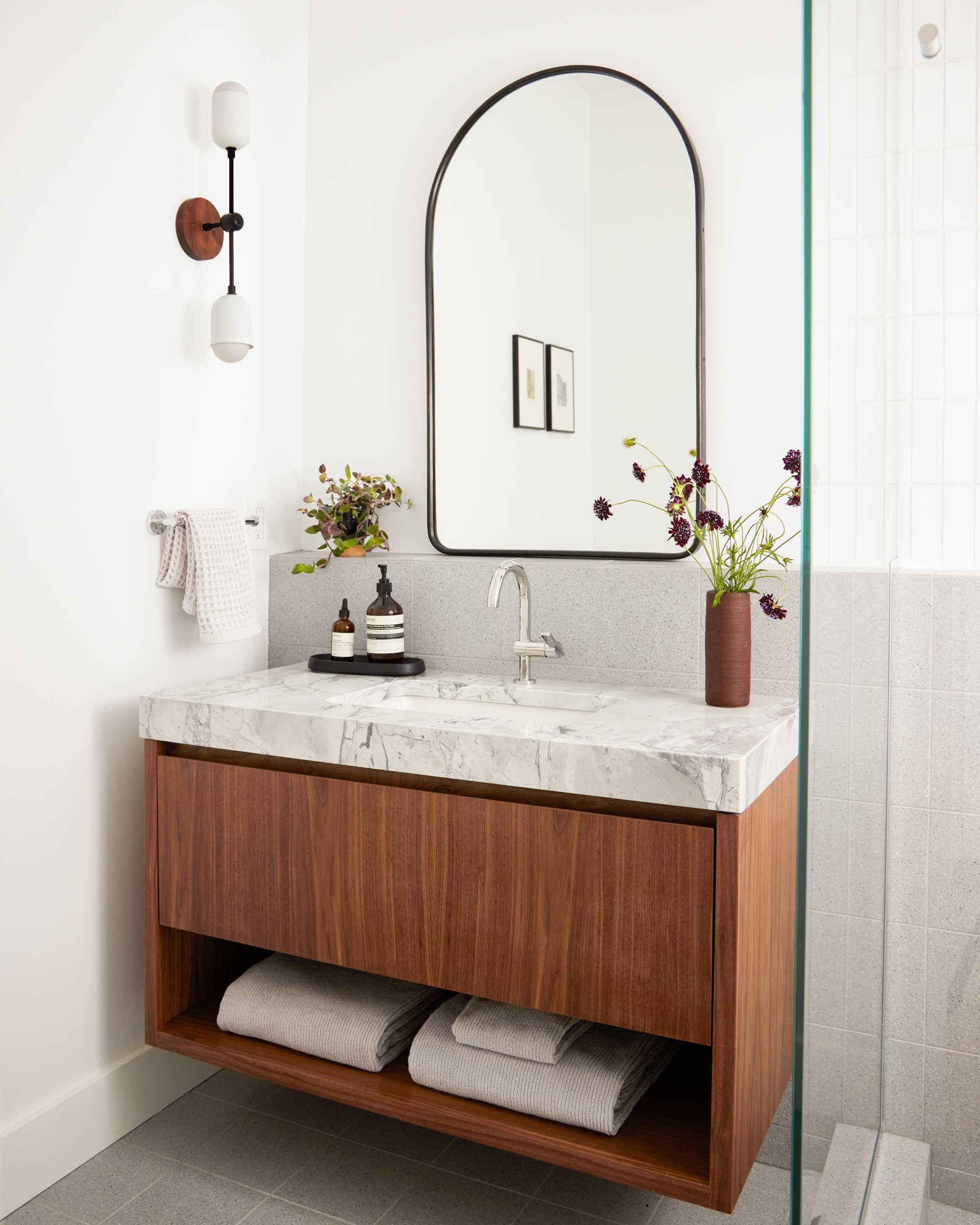 A remodeled bathroom includes a new wood vanity with a large drawer and a shelf below is topped with a thick countertop with undermount sink, while a ledge below the mirror carries through to the shower.