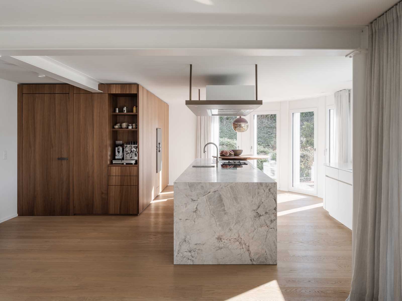 A modern kitchen with wood cabinets, wood floors, and a long kitchen island with a stone waterfall countertop.