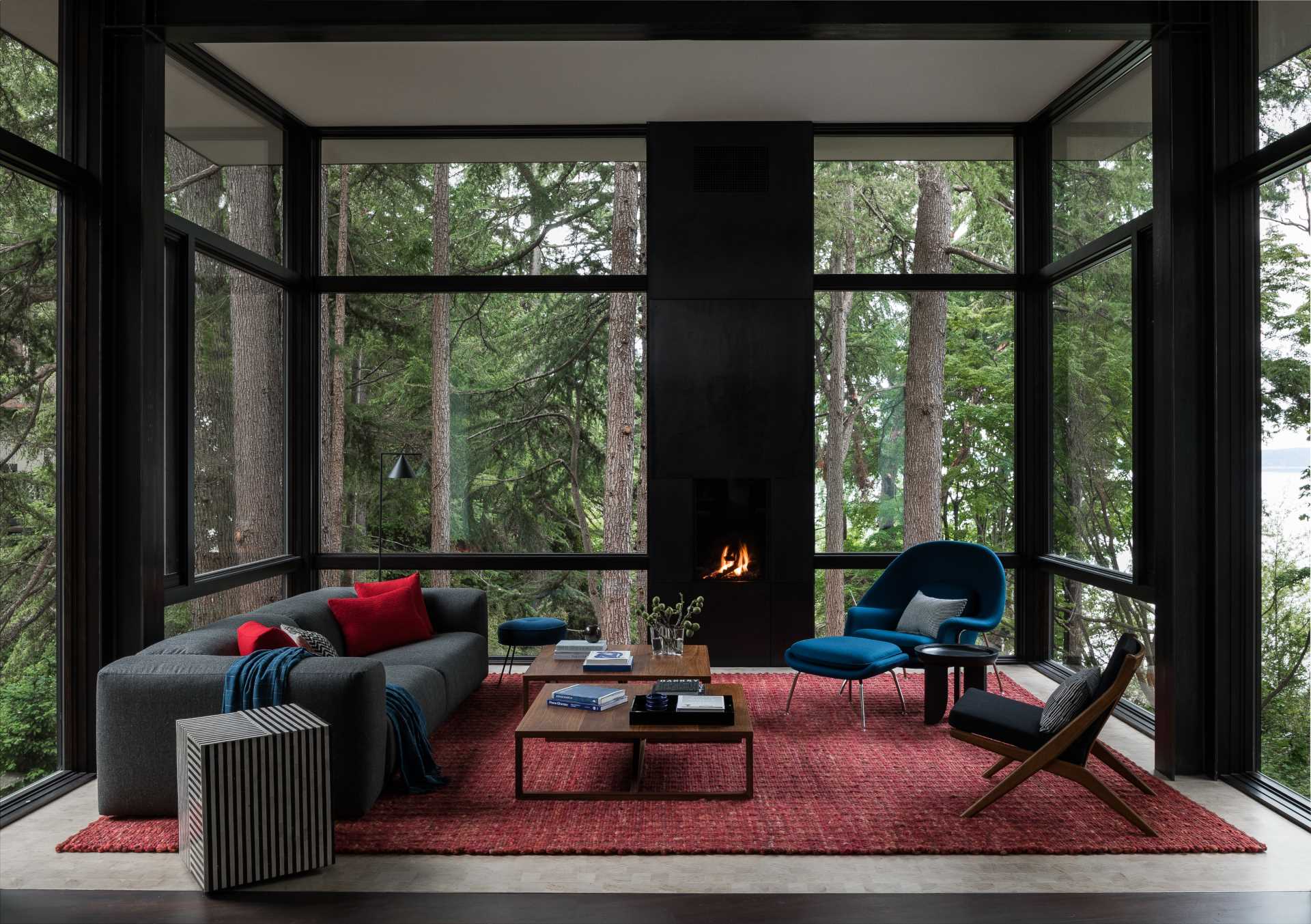 This modern living room, which is surrounded by trees on three sides, includes thick black window frames and a fireplace, while a bold reg rug and blue chair add a bold splash of color to the space.