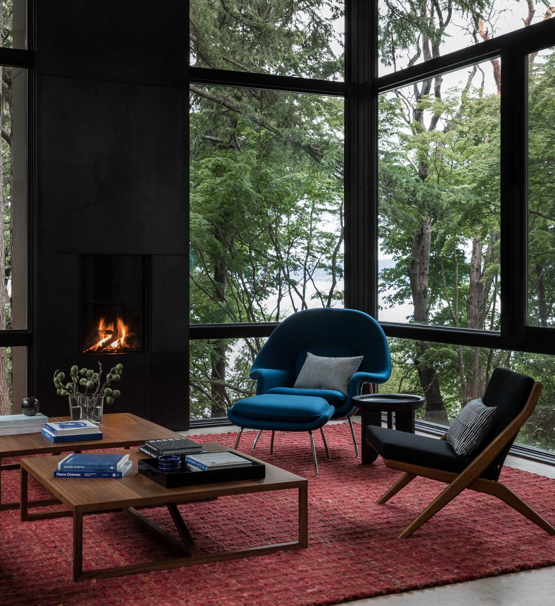 This modern living room, which is surrounded by trees on three sides, includes thick black window frames and a fireplace, while a bold reg rug and blue chair add a bold splash of color to the space.