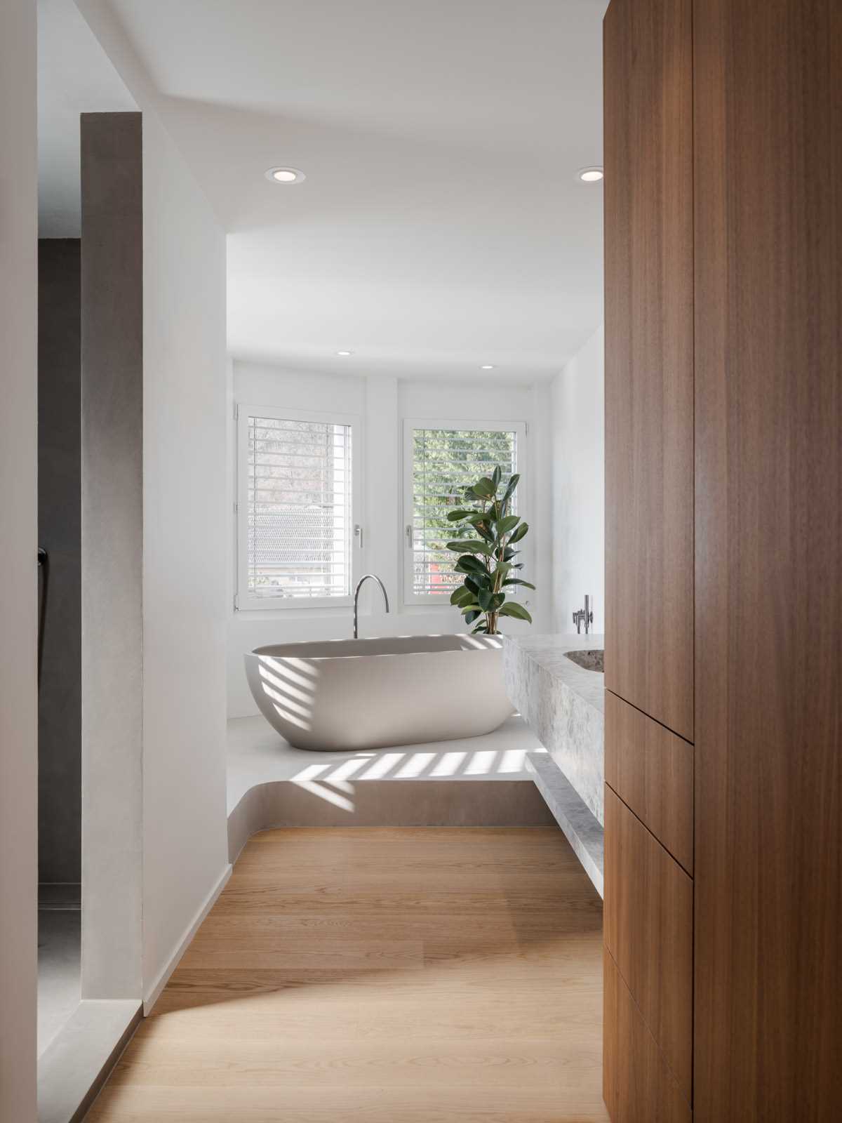 This modern primary bathroom features a freestanding bathtub raised up on a platform by the window, while the stone vanity has a built-in sink and adjacent tall wood cabinet.