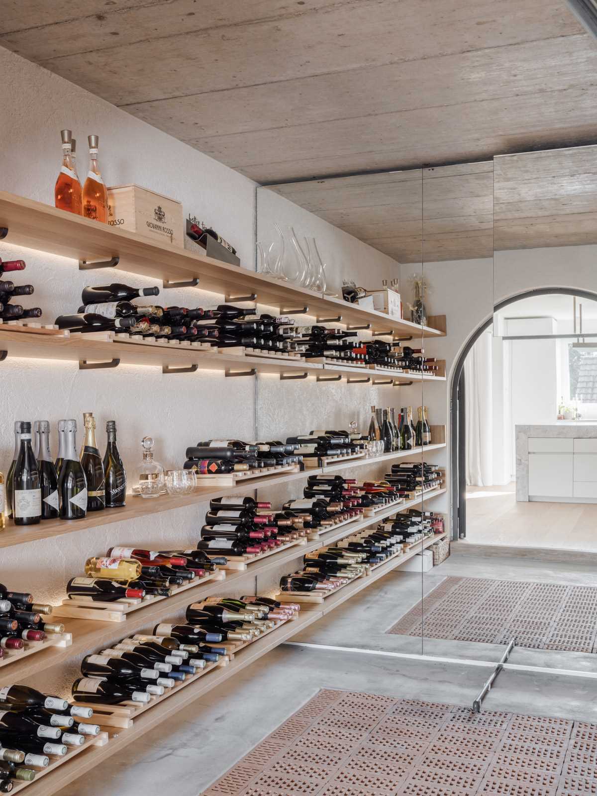 An arched glass doorway with a thick black frame provides a glimpse of this modern wine cellar, where a mirror makes the cellar seem much larger than it is, and wood shelves with hidden lighting provide plenty of wine storage.