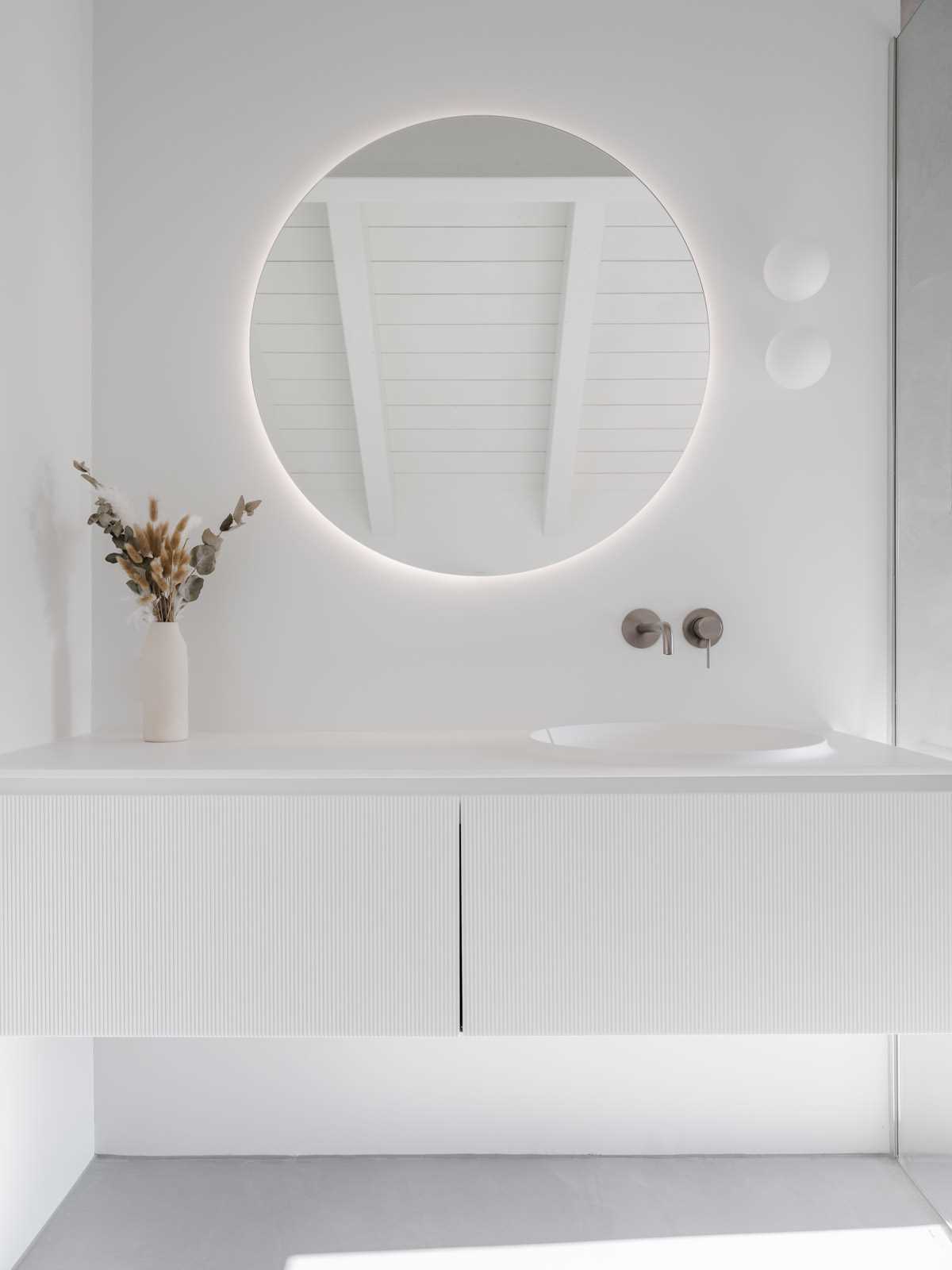 In this modern bathroom, there's a round backlit mirror mounted on the wall above a white vanity with a textured front. Next to the vanity area is the shower which includes a shelving niche.