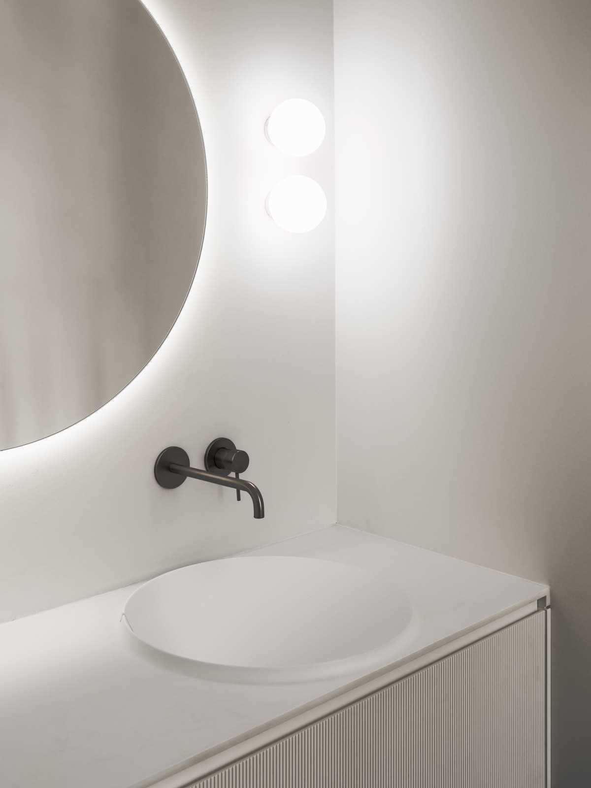 In this modern bathroom, there's a round backlit mirror mounted on the wall above a white vanity with a textured front. Next to the vanity area is the shower which includes a shelving niche.