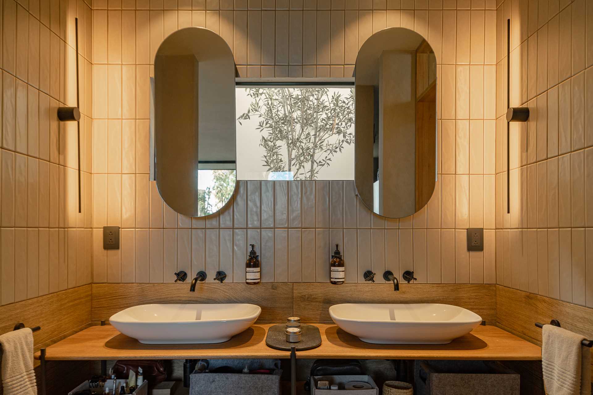یک حمام مدرن با روشویی دو نفره، یک پنجره کوچک، آینه های قرصی شکل، کاشی دیواری که تا سقف می رود، و یک دوش با کاشی تیره تر.