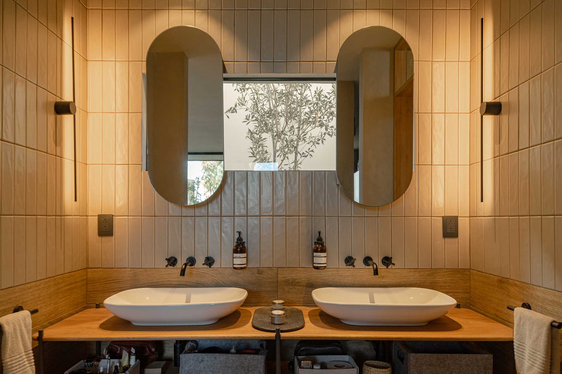 A modern bathroom with double vanity, a small window, pill-shaped mirrors, wall tile that goes to the ceiling, and a walk-in shower with a darker tile.