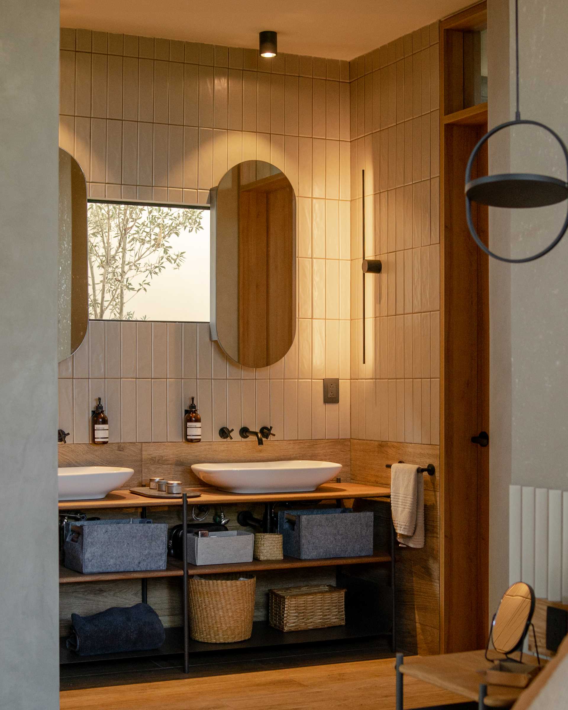 A modern bathroom with double vanity, a small window, pill-shaped mirrors, wall tile that goes to the ceiling, and a walk-in shower with a darker tile.