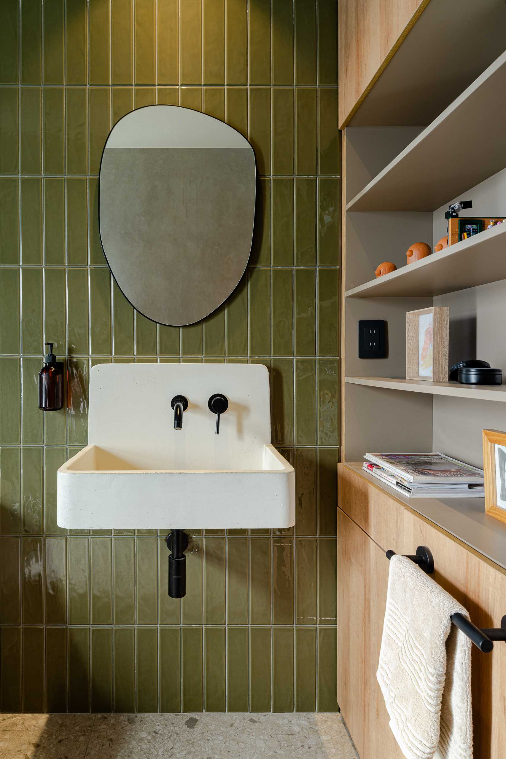 A modern bathroom with vertical green wall tiles, a white basin, and a built-in wood storage unit with open shelves.