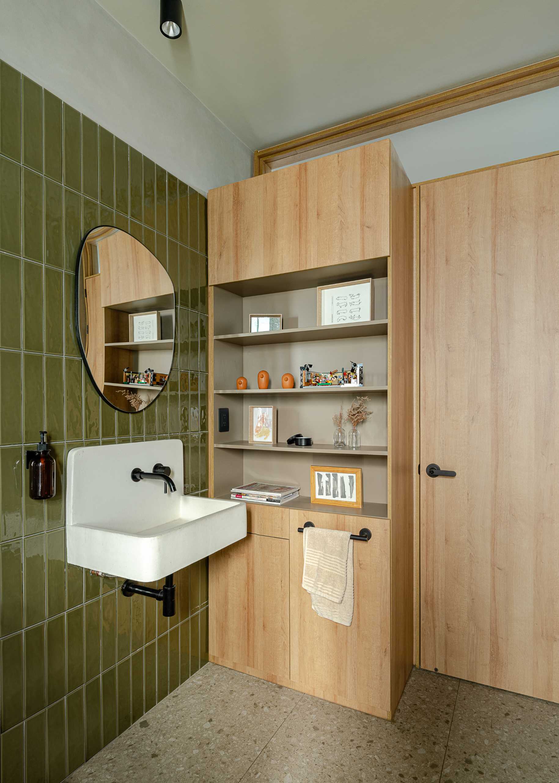 A modern bathroom with vertical green wall tiles, a white basin, and a built-in wood storage unit with open shelves.