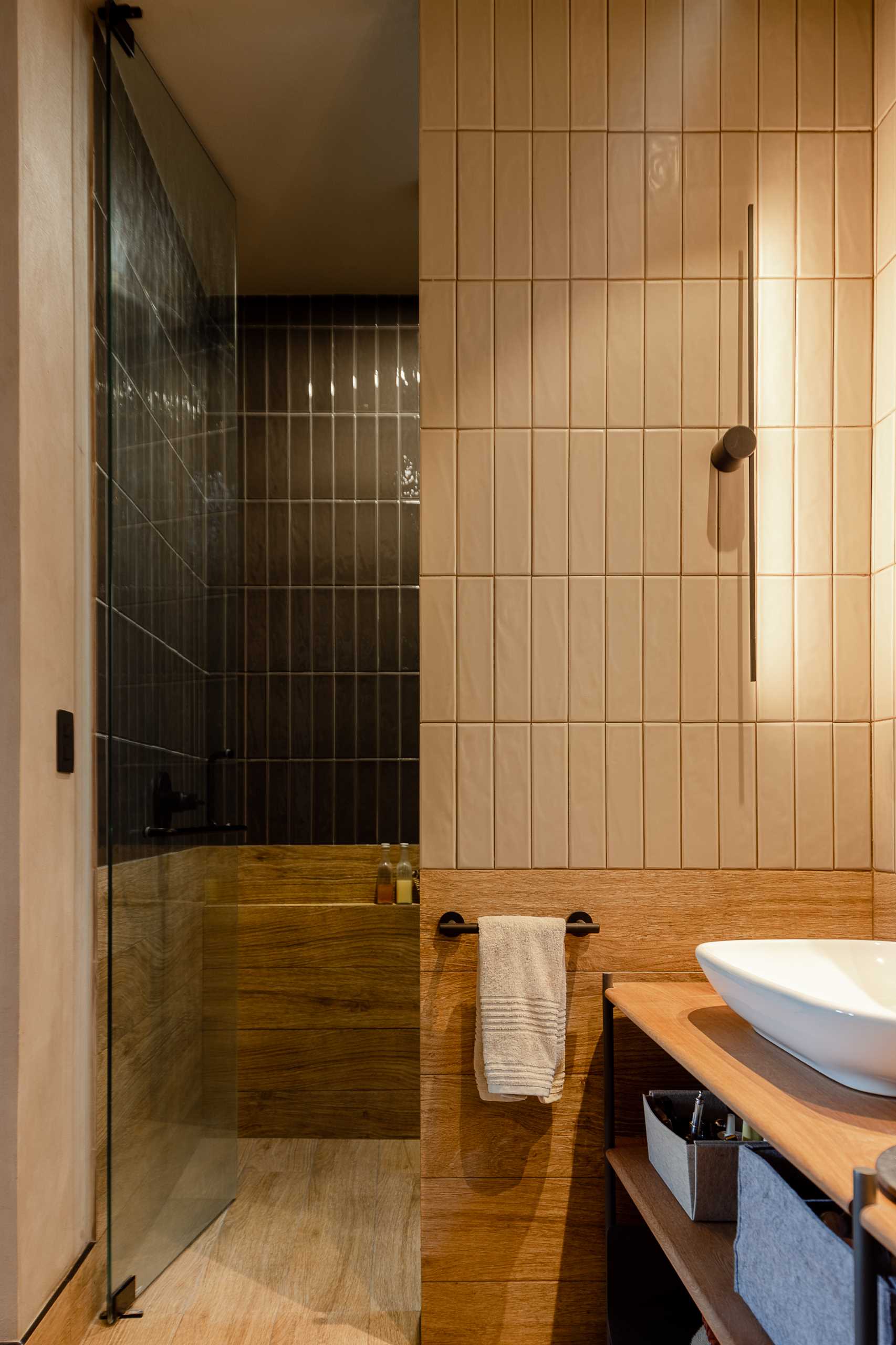 A modern bathroom with double vanity, a small window, pill-shaped mirrors, wall tile that goes to the ceiling, and a walk-in shower with a darker tile.