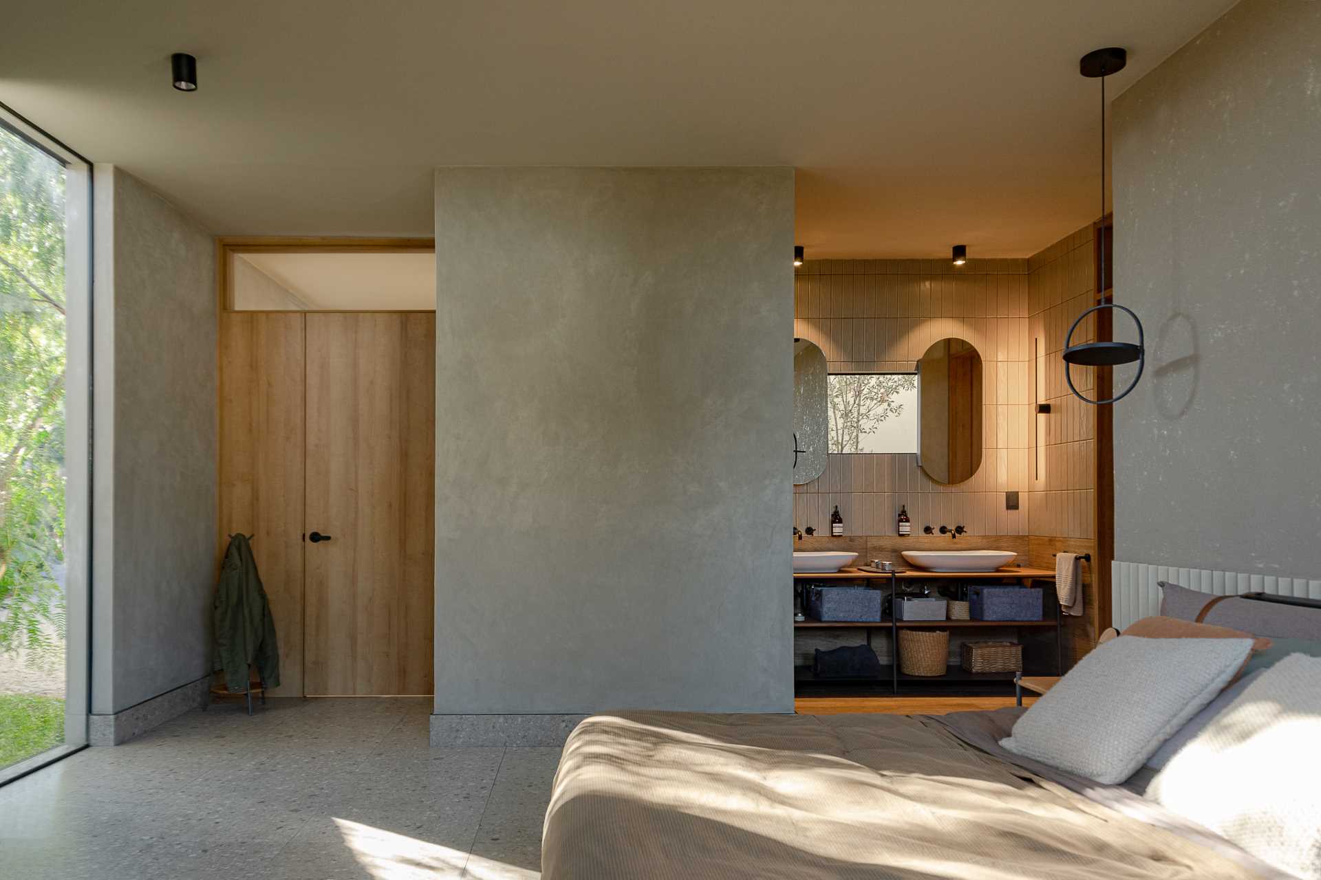 A modern bathroom with double vanity, a small window, pill-shaped mirrors, wall tile that goes to the ceiling, and a walk-in shower with a darker tile.