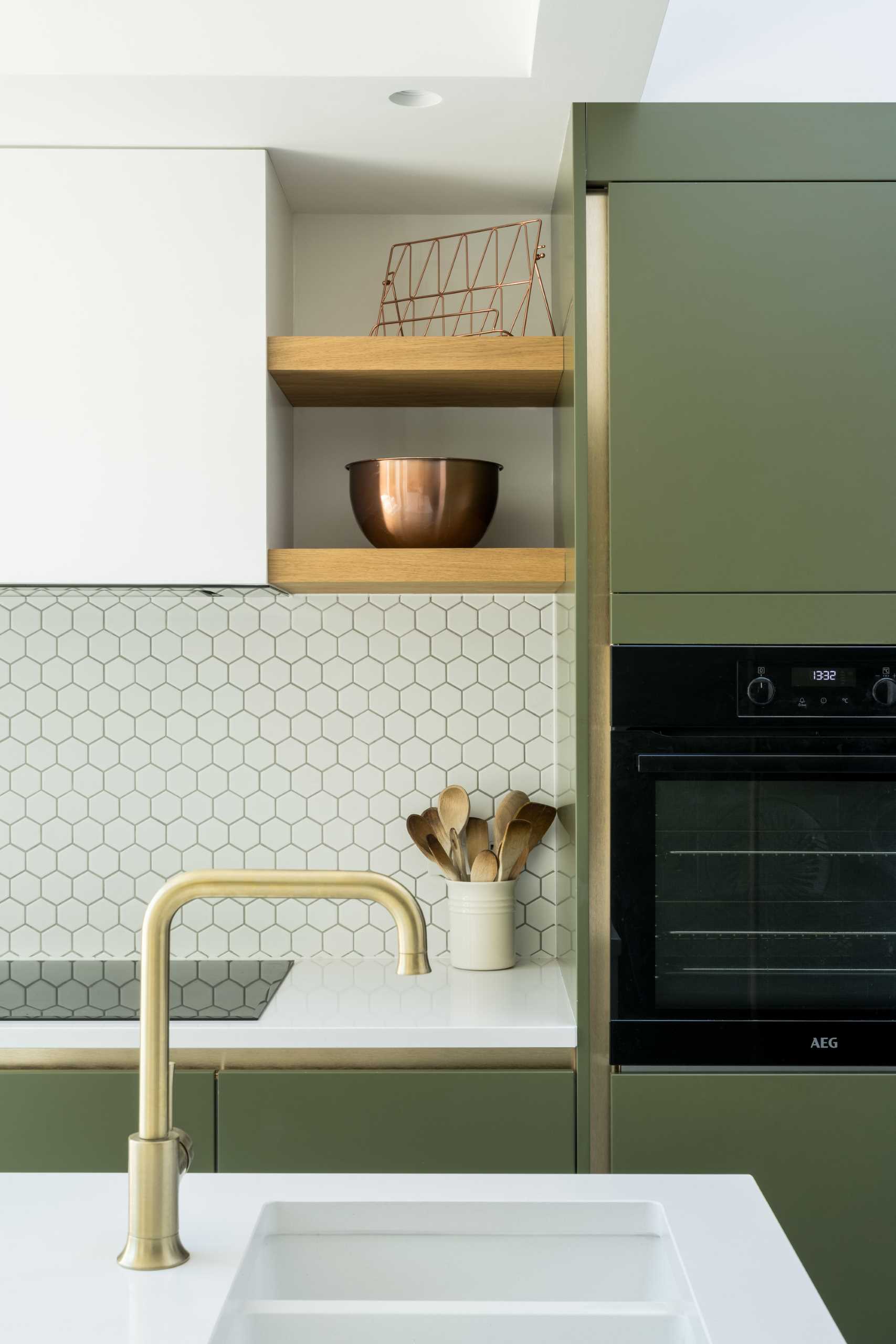 The new kitchen includes matte green cabinets, small hexagonal white tile backsplashes, wood shelves, and an island with undermount sink.