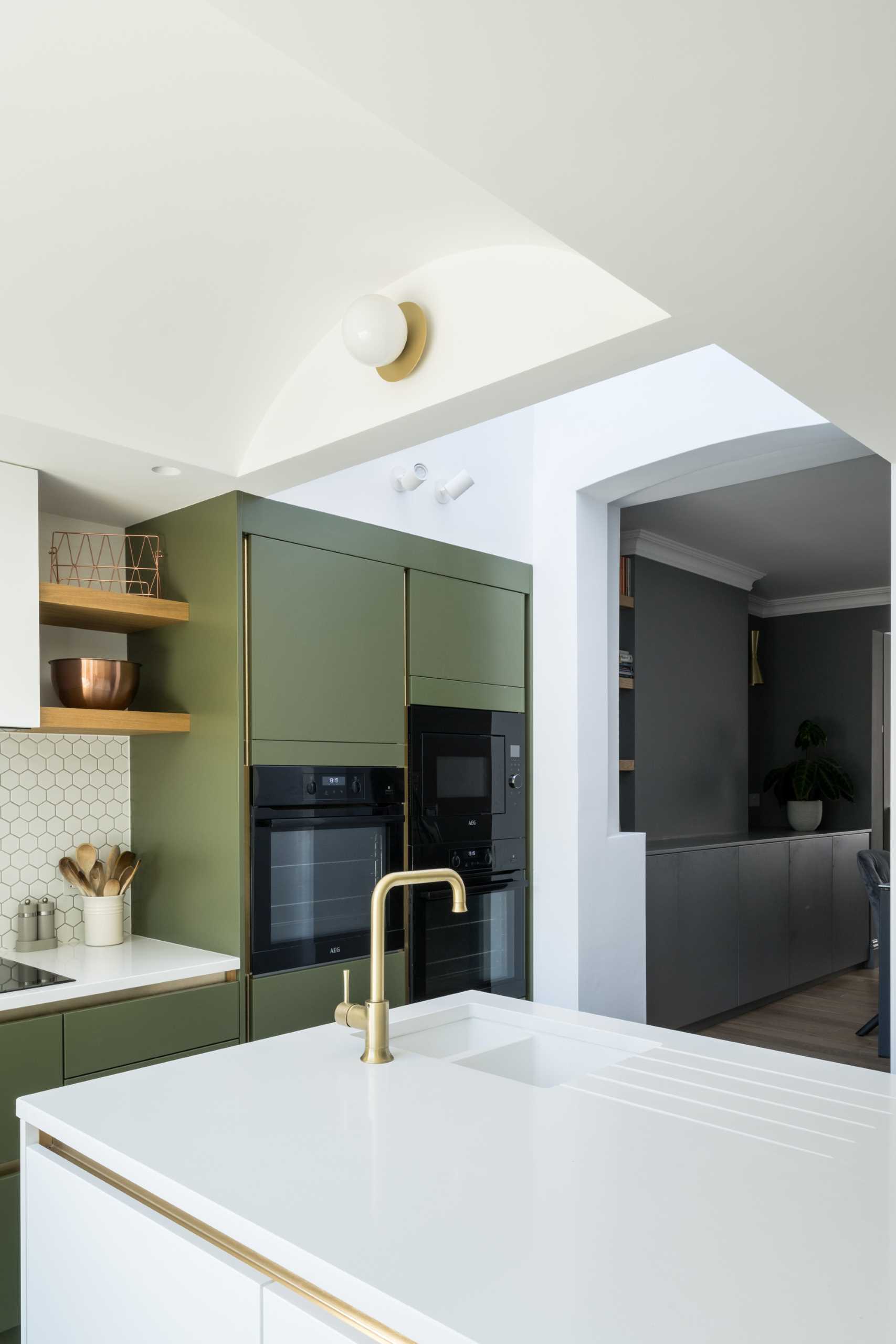 The new kitchen includes matte green cabinets, small hexagonal white tile backsplashes, wood shelves, and an island with undermount sink.