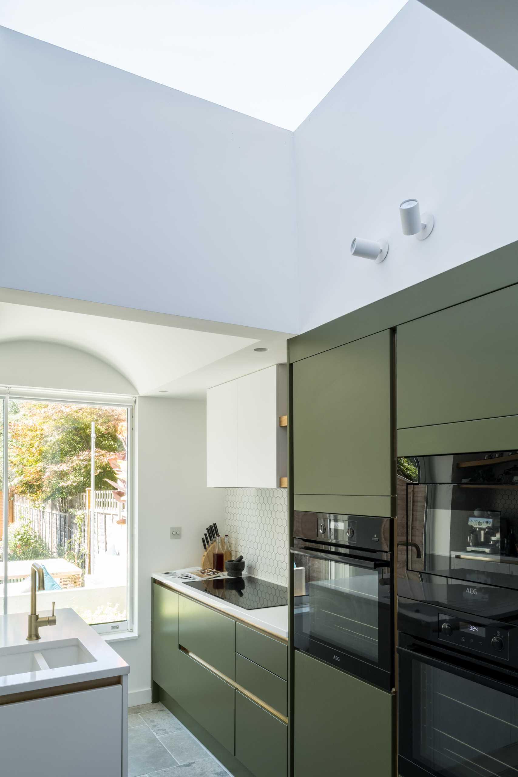 The new kitchen includes matte green cabinets, small hexagonal white tile backsplashes, wood shelves, and an island with undermount sink.