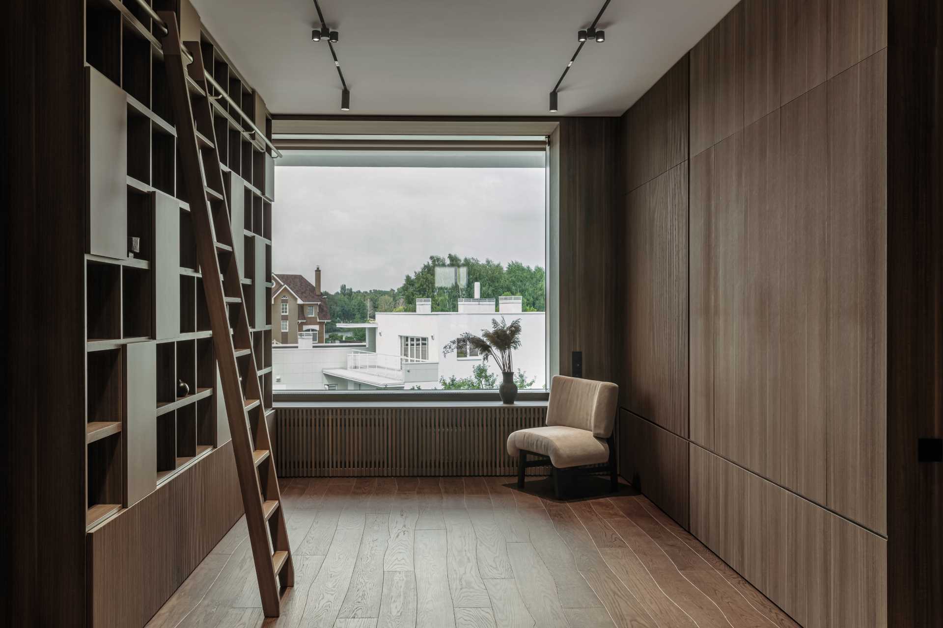 A library with a large window looking out onto the neighborhood, while wood walls, shelving, and flooring add warmth to the space.