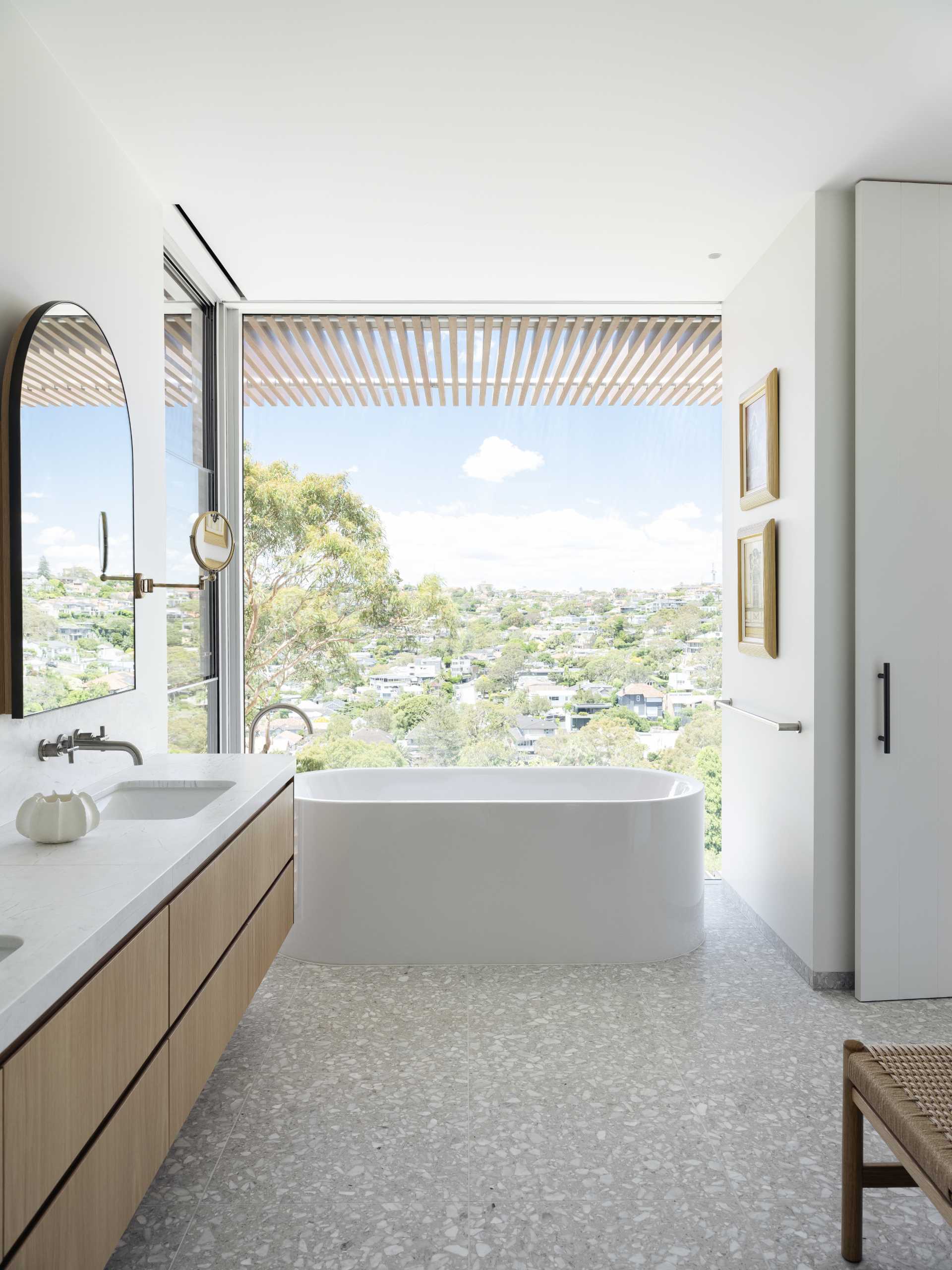 In this bathroom, a large picture window provides a backdrop for the freestanding bathtub, and arched mirrors are hung on the wall above the double vanity. 