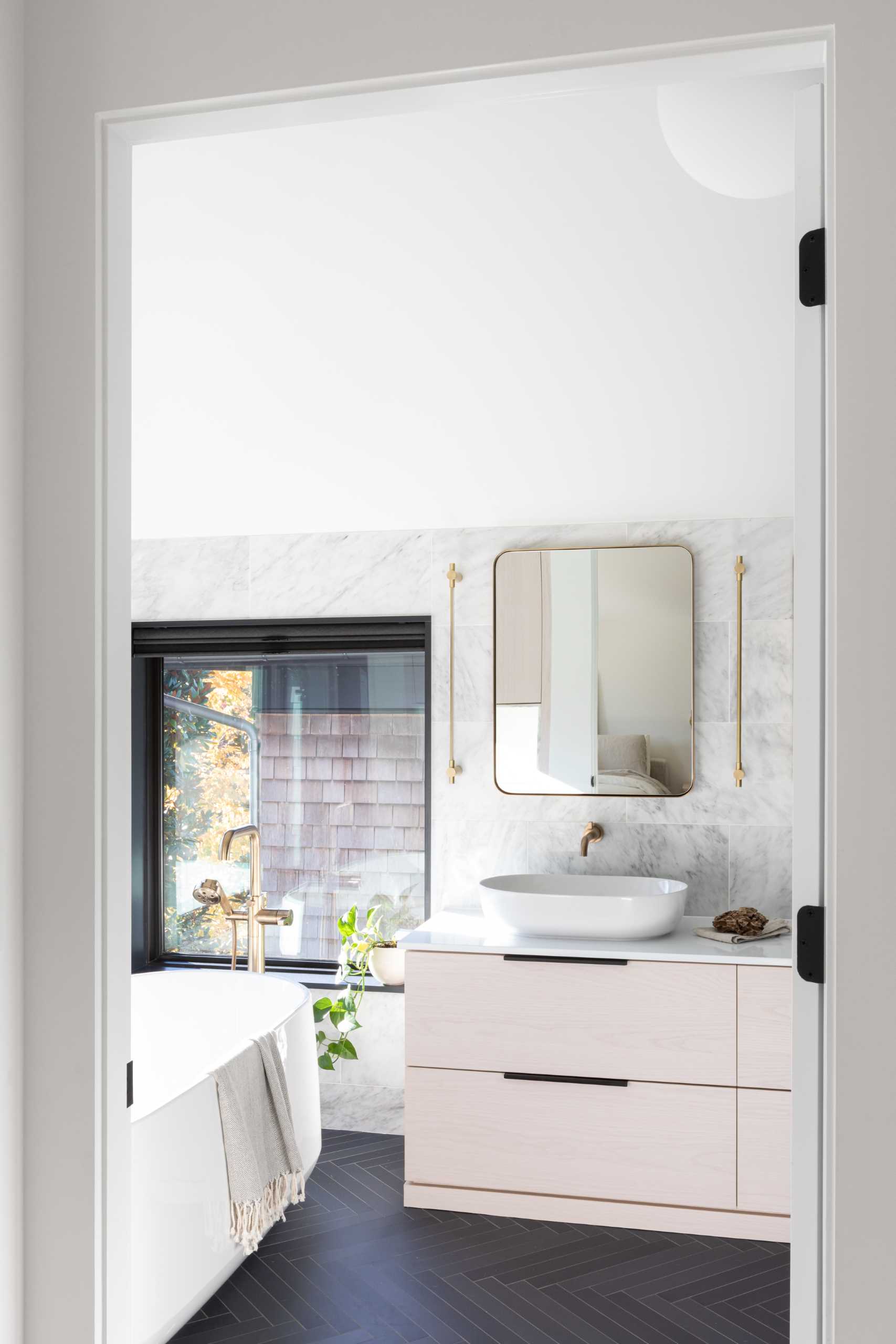 In this modern bathroom, there's wood cabinetry, a freestanding bathtub, dark floor tiles in a herringbone pattern, and metallic accents.