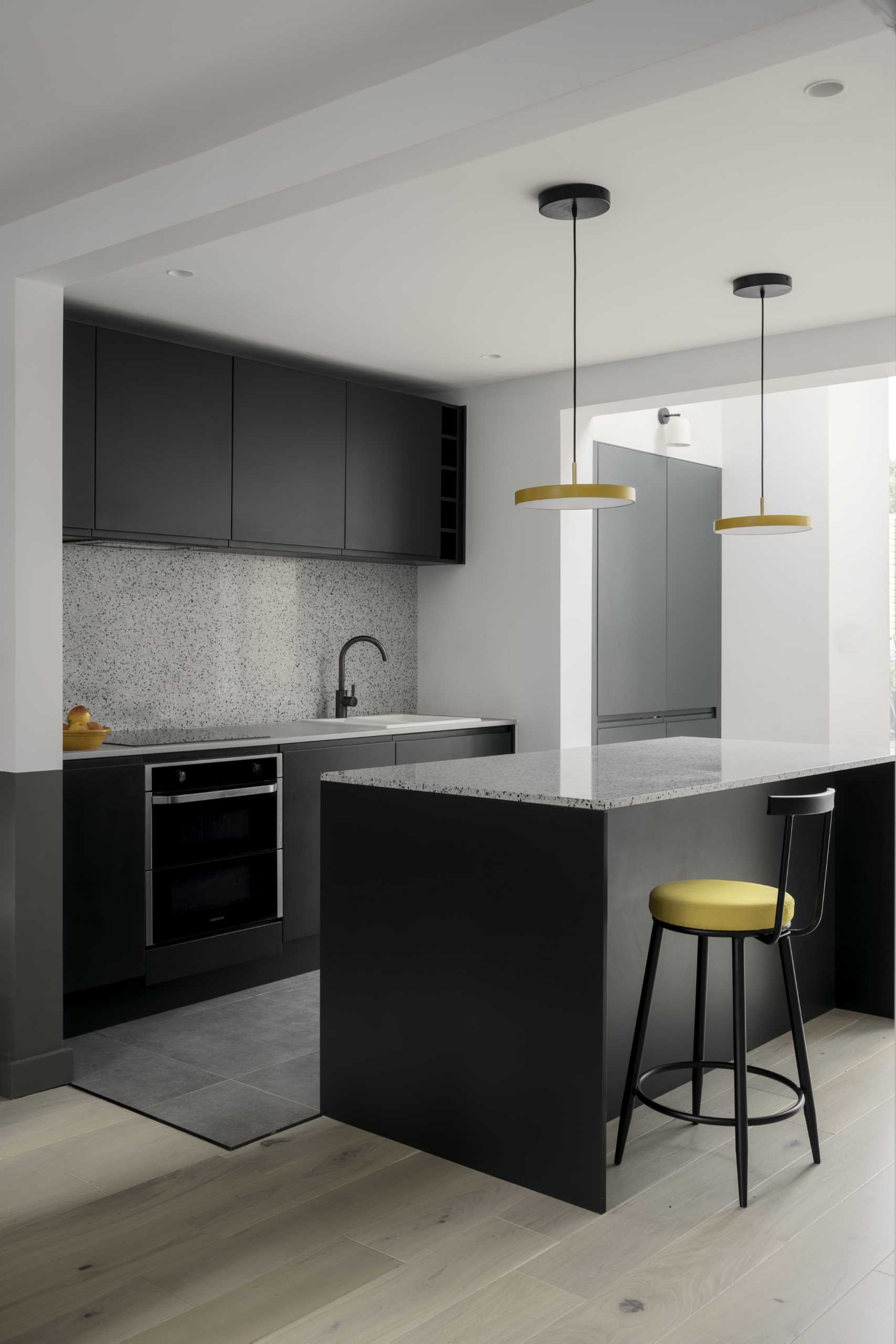 A kitchen with minimalist black cabinets and an island with seating. Yellow accents in the lights and chairs add a pop of color to the space.
