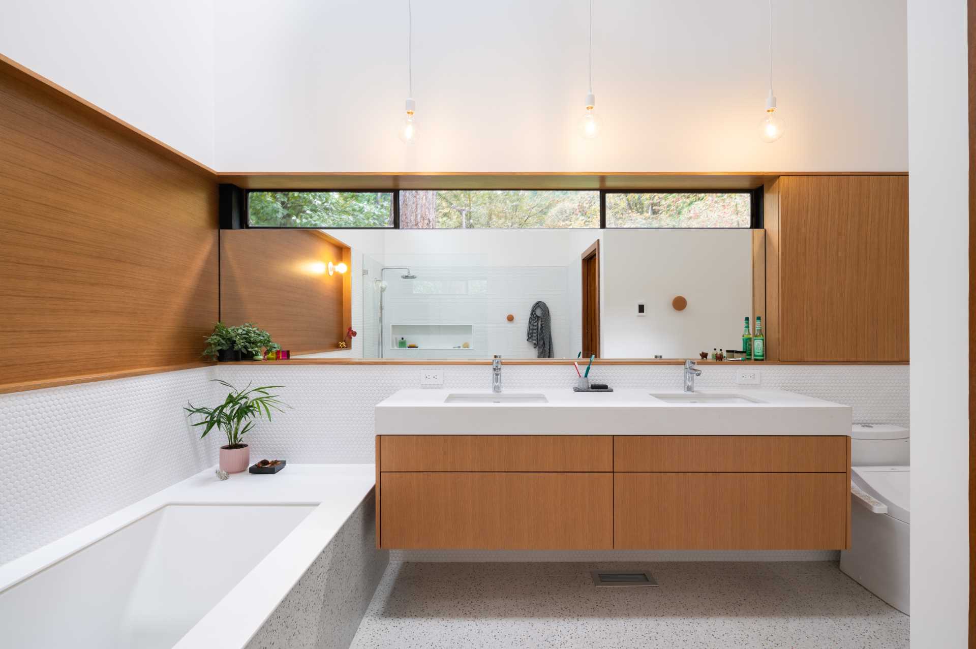 یک حمام مدرن با دیوارهای پوشیده از کاشی‌های پنی سفید، یک وان حمام توکار، دوش با طاقچه قفسه‌بندی، و جزئیات چوبی پیچیده‌شده برای مطابقت با روشویی دوتایی.