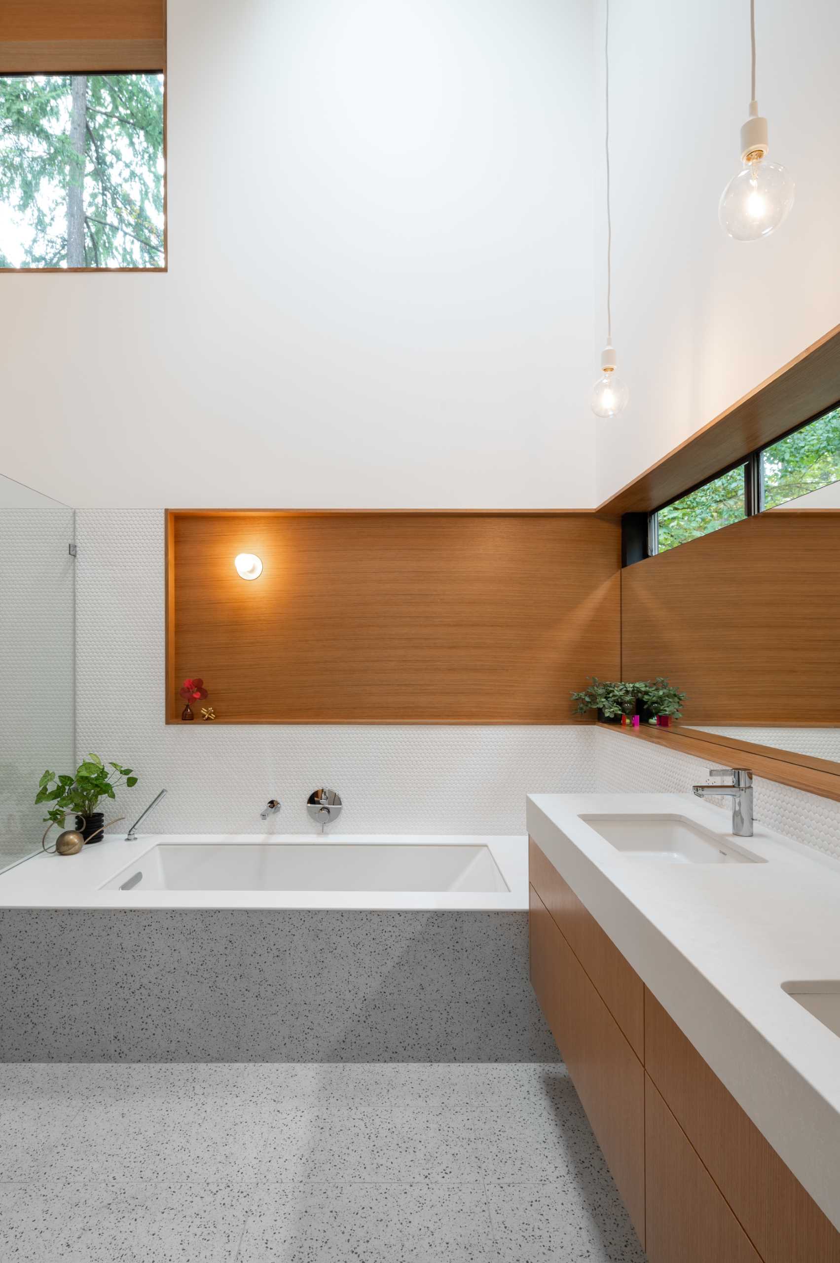 A modern bathroom with walls covered in white penny tiles, a built-in bathtub, a walk-in shower with a shelving niche, and a wrap-around wood detail to match the double vanity.