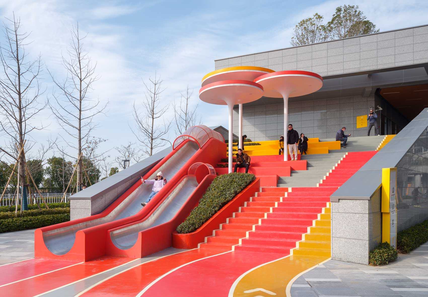 A modern and colorful landscaped park with slides, pathways, a merry-go-round, lounge seating, seesaw, shading structures, seating, swings, and an amphitheater.