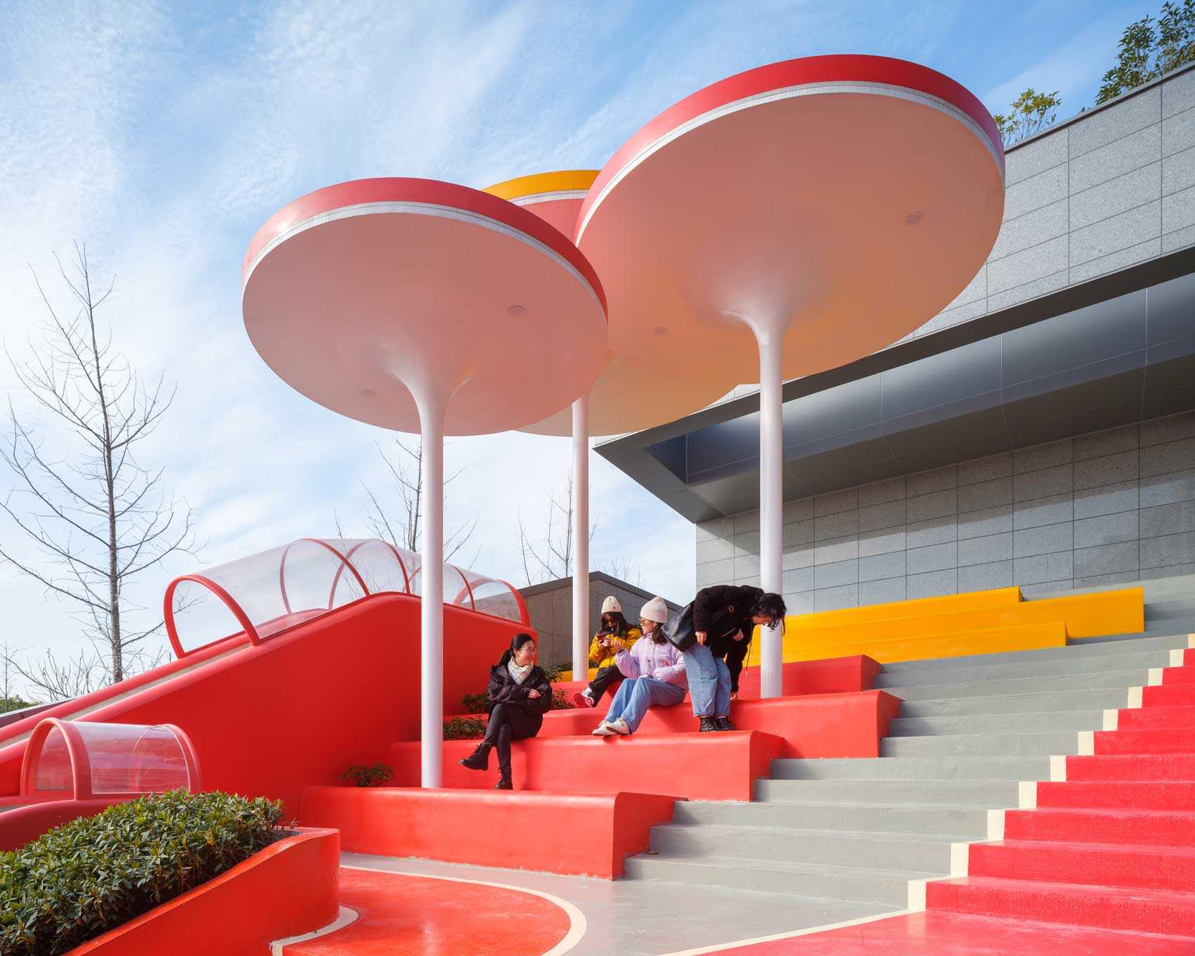 A modern and colorful landscaped park with slides, pathways, a merry-go-round, lounge seating, seesaw, shading structures, seating, swings, and an amphitheater.