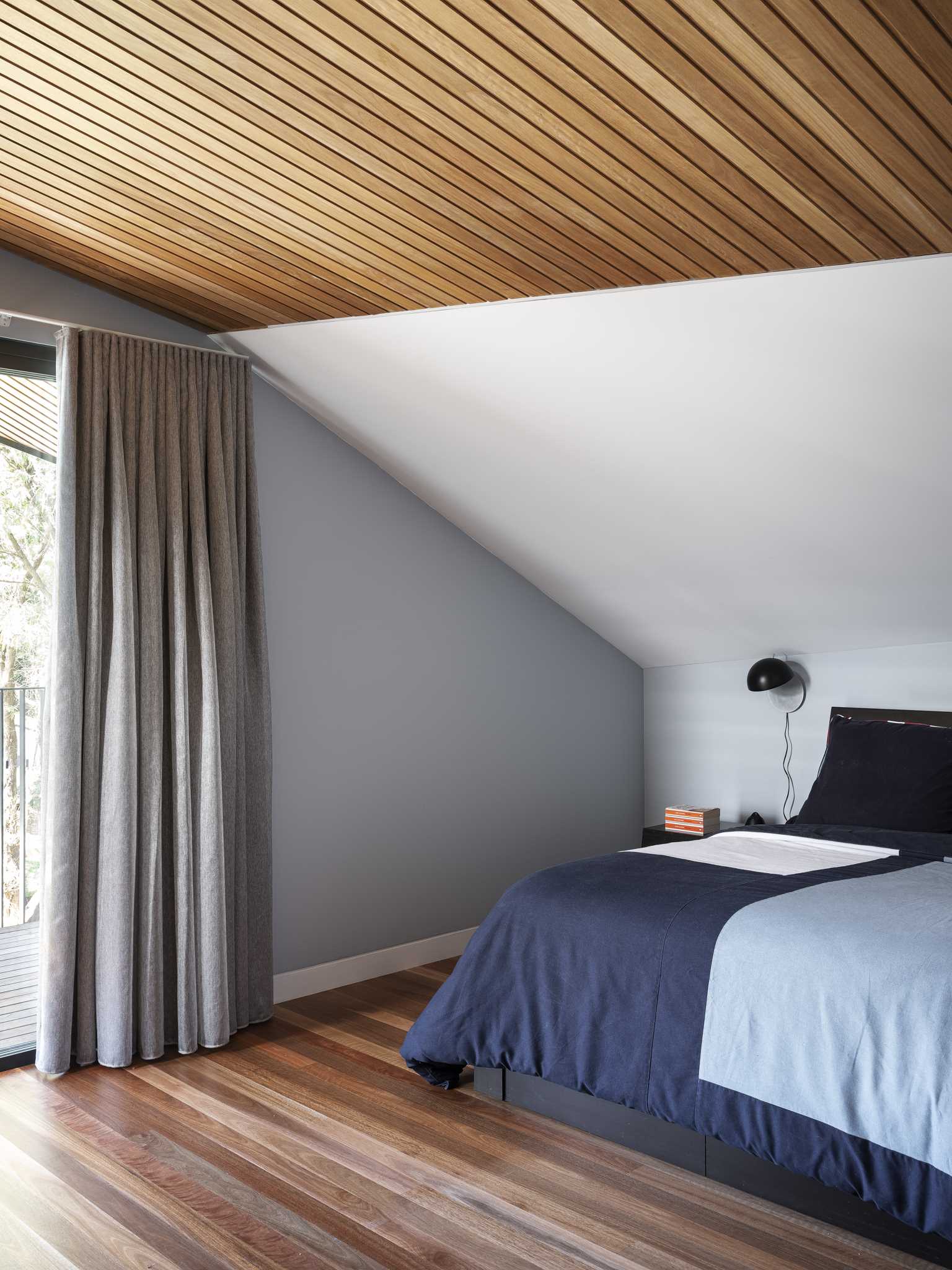 In this modern bedroom, there's a wood ceiling and a wood floor, while a grey accent wall surrounds a sliding glass door that opens to a balcony.
