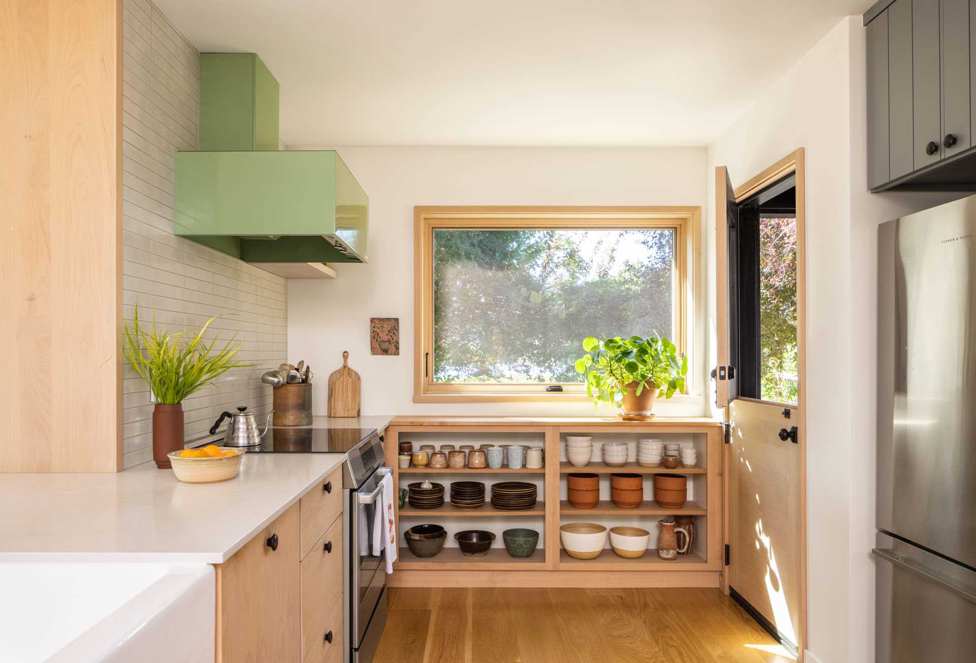 A remodeled kitchen with new light wood cabinets and open shelving, a pastel green range hood, a light-tiled wall, and wood floors. A Dutch door was also added.